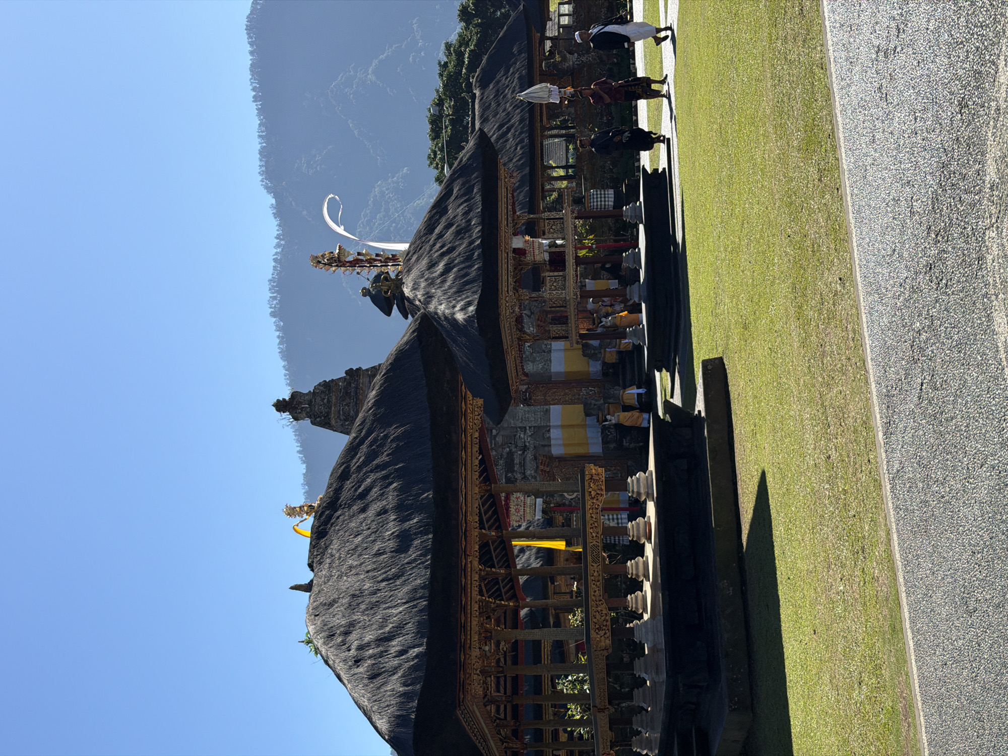 The image depicts a clear, sunny day at Pura Ulun Danu Batur temple in Baturiti, Indonesia. Several traditional Balinese structures with thatched roofs and intricate wooden carvings are visible. In the foreground, a well-maintained grassy area is separated from a gravel path by a curb.
Several people are present within the temple grounds. On the right side of the image, three men are walking across the lawn. The man in the foreground is wearing a white shirt and pants with a black jacket. The man behind him is wearing a dark outfit and a sarong. The furthest man is wearing a dark shirt and a sarong, and is carrying a white umbrella-like object on his head.
In the middle ground, two figures in yellow attire are standing near one of the structures. Another figure in yellow is visible further back within the structure. A yellow flag hangs from the roof of the structure on the left.
The background features more temple structures and lush green trees. Beyond the trees, a majestic mountain range rises against a clear blue sky. The overall scene conveys a sense of cultural and natural beauty.
