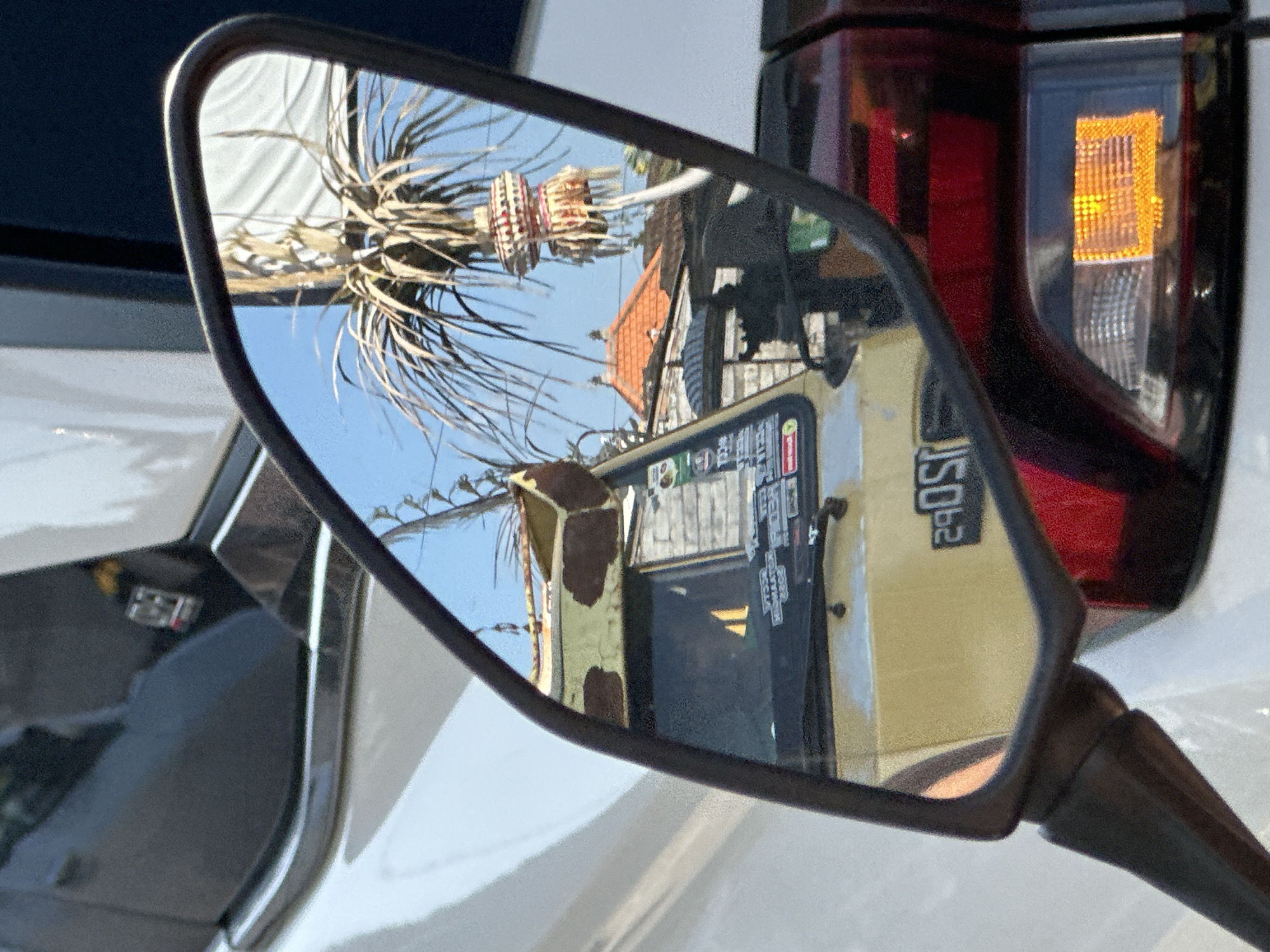 The image is a close-up, side-angle shot of a vehicle's side mirror, reflecting a scene in North Kuta, Indonesia. The mirror is mounted on a white vehicle. The reflection shows a yellow truck with a damaged, rusted top section that is painted with irregular brown patches. The truck's windshield has several stickers. Behind the truck, traditional Balinese roof tiles in orange are visible. A tall palm tree with dried fronds stands prominently, and an ornate, multi-tiered decorative item, possibly a canang sari offering, hangs from its branches. The sky in the reflection is a clear blue. The truck's license plate, partially visible in the reflection and reversed, appears to read "2905". The overall scene suggests a street or roadside environment in a tropical setting, likely populated by both vehicles and cultural elements.