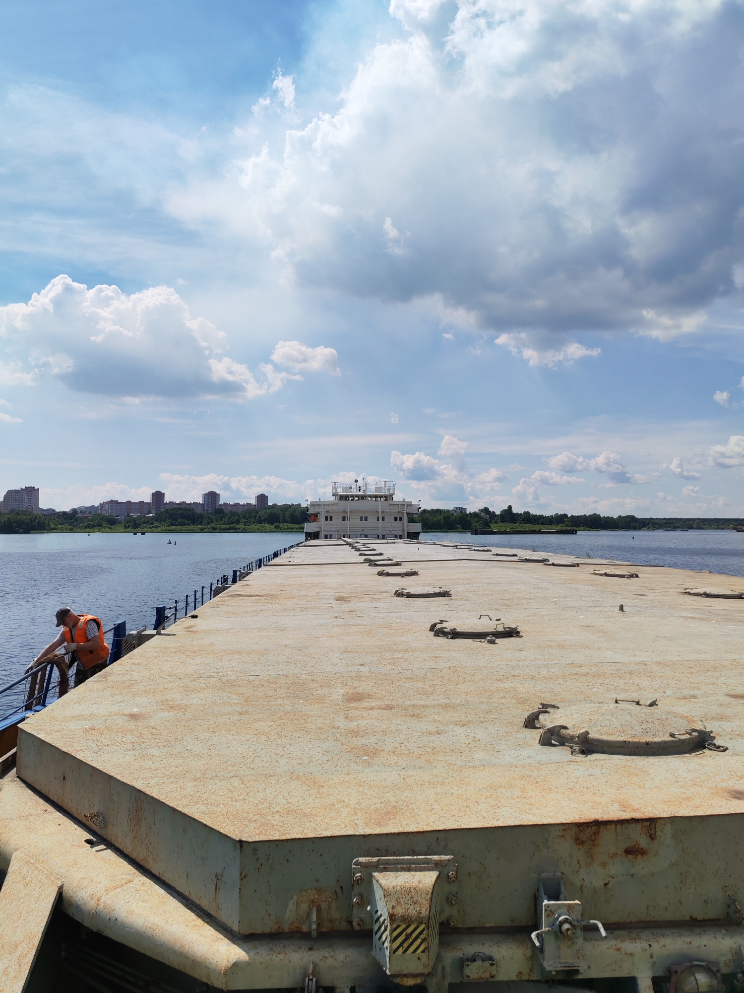This image depicts a view from the deck of a barge, possibly a cargo barge, on a river. A man wearing an orange safety vest and a cap is leaning over a railing on the left side of the barge, holding onto a rope. In the background, across the river, is the cityscape of Cherepovets, Russia, with several multi-story buildings visible. The sky is partly cloudy with dramatic cumulus clouds. The deck of the barge is concrete and shows signs of wear, with several circular hatches or manholes. A white superstructure building is visible at the far end of the barge. The water appears calm and blue. The scene suggests a working day or operational status of the barge.