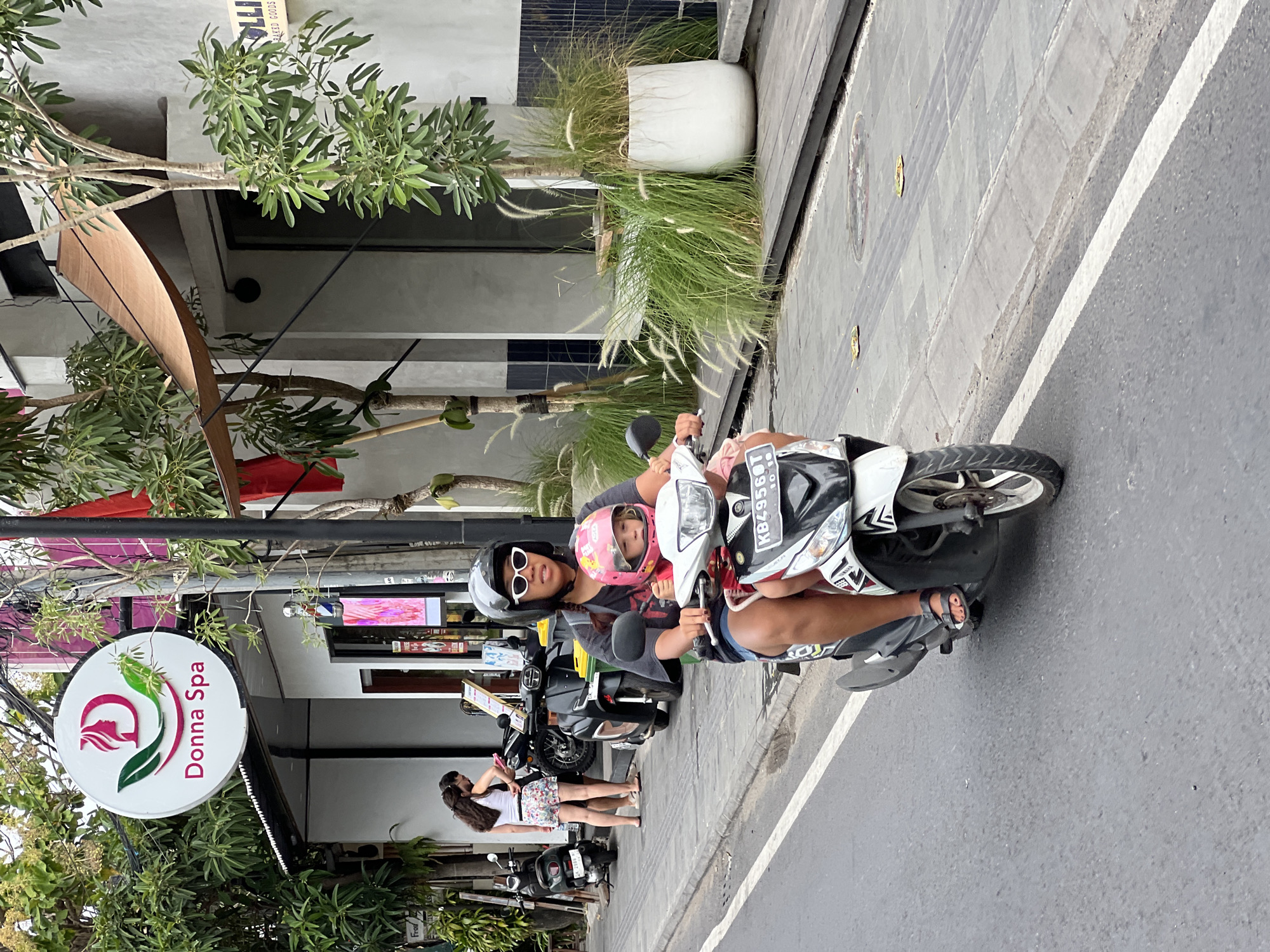In Denpasar, Indonesia, a woman wearing a dark t-shirt, denim shorts, a helmet, and white sunglasses rides a scooter. A young child in a pink helmet sits in front of her. The scooter is white and black, with a license plate that reads "KB4956BT 11-25". They are on a paved road with a white line marking the lane. To the right of the road, there is a raised wooden walkway with potted plants and trees. In the background, a sign for "Donna Spa" is visible, featuring a pink outline of a woman's profile and two green leaves. Other motorcycles are parked along the sidewalk.