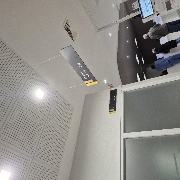 This image depicts an interior scene within a medical facility, specifically a radiology department, located in Strasbourg, France. The overhead area features a light-colored suspended ceiling constructed from perforated acoustic tiles, interspersed with square recessed LED light fixtures and a rectangular ventilation grille. Two black directional signs with yellow accents are visible: a suspended sign prominently displays "Attente Scanner - IRM" (Waiting Scanner - MRI), and a wall-mounted sign with a left-pointing arrow reads "← RM - Radiologie" (Radiology). On the left side, two large panels of frosted glass are visible, likely forming part of a door or partition. To the right, three individuals are positioned at a white reception counter. One person wears blue medical scrubs, standing with their back to the camera. Adjacent to them, a second individual with grey hair, dressed in a dark jacket, also faces the counter. A third person, wearing a grey patterned jacket and blue jeans, stands with their back to the camera, facing the counter area. A white digital self-service kiosk with a blue screen interface is situated to the right of the counter.