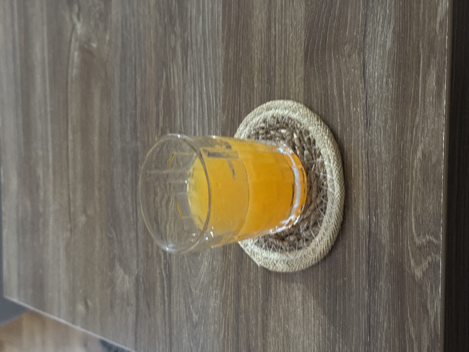 The scene, located in Cibukovac, Serbia, displays a clear glass containing a vibrant orange liquid. The glass, approximately two-thirds full, features a distinct geometric pattern around its upper circumference. It rests centrally on a round coaster constructed from woven natural fibers with a braided outer edge. Both objects are positioned on a light brown surface with a visible wood grain pattern, consistent with a laminate or wooden table. The background is out of focus, showing a muted, blurred grey area in the upper left quadrant. No individuals, explicit actions, or direct interactions are present.