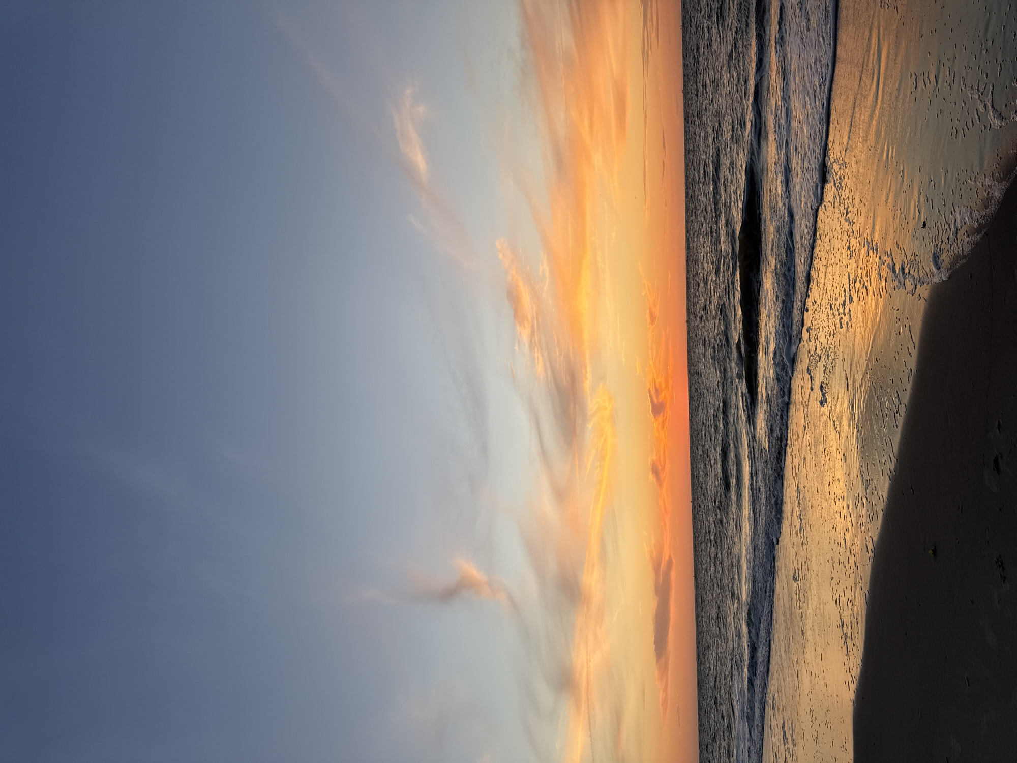 A wide-angle vertical shot captures a serene sunset over the North Sea beach on Langeoog, Germany. The sky transitions from a clear, pale blue at the top to vibrant hues of orange and yellow closer to the horizon, where thin, wispy clouds are illuminated by the setting sun. The ocean is a dark, choppy expanse with gentle waves lapping at the shore. The wet sand reflects the warm colors of the sky, creating a shimmering effect. The foreground consists of dark, damp sand. No people or animals are visible in the frame.