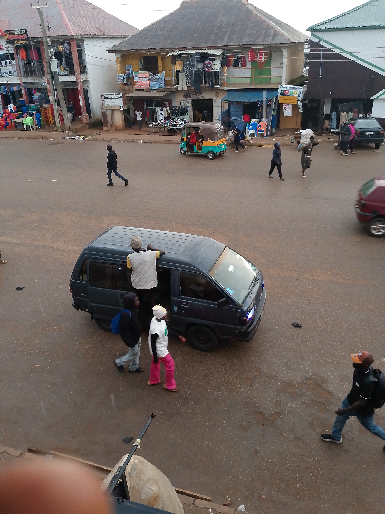 In Bukuru, Nigeria, a busy street scene unfolds. A dark blue minivan, its rear door open, is the central focus. A man wearing a beige hat and a yellow t-shirt leans over the minivan's roof, seemingly assisting or observing. Beside the open door, a person in a black hoodie and a blue backpack stands, while another individual in a white t-shirt and bright pink pants is positioned next to them. Further down the street, a man in a black jacket and blue jeans walks away from the minivan, and another man wearing a black shirt, a brown and orange cap, a backpack, and blue jeans walks towards the right of the frame. In the background, various shops line the street, with some displaying merchandise like clothes and footwear. A three-wheeled auto-rickshaw is parked on the street. The road surface appears wet, suggesting recent rain. The overall atmosphere is one of daily commerce and pedestrian activity in a Nigerian town.