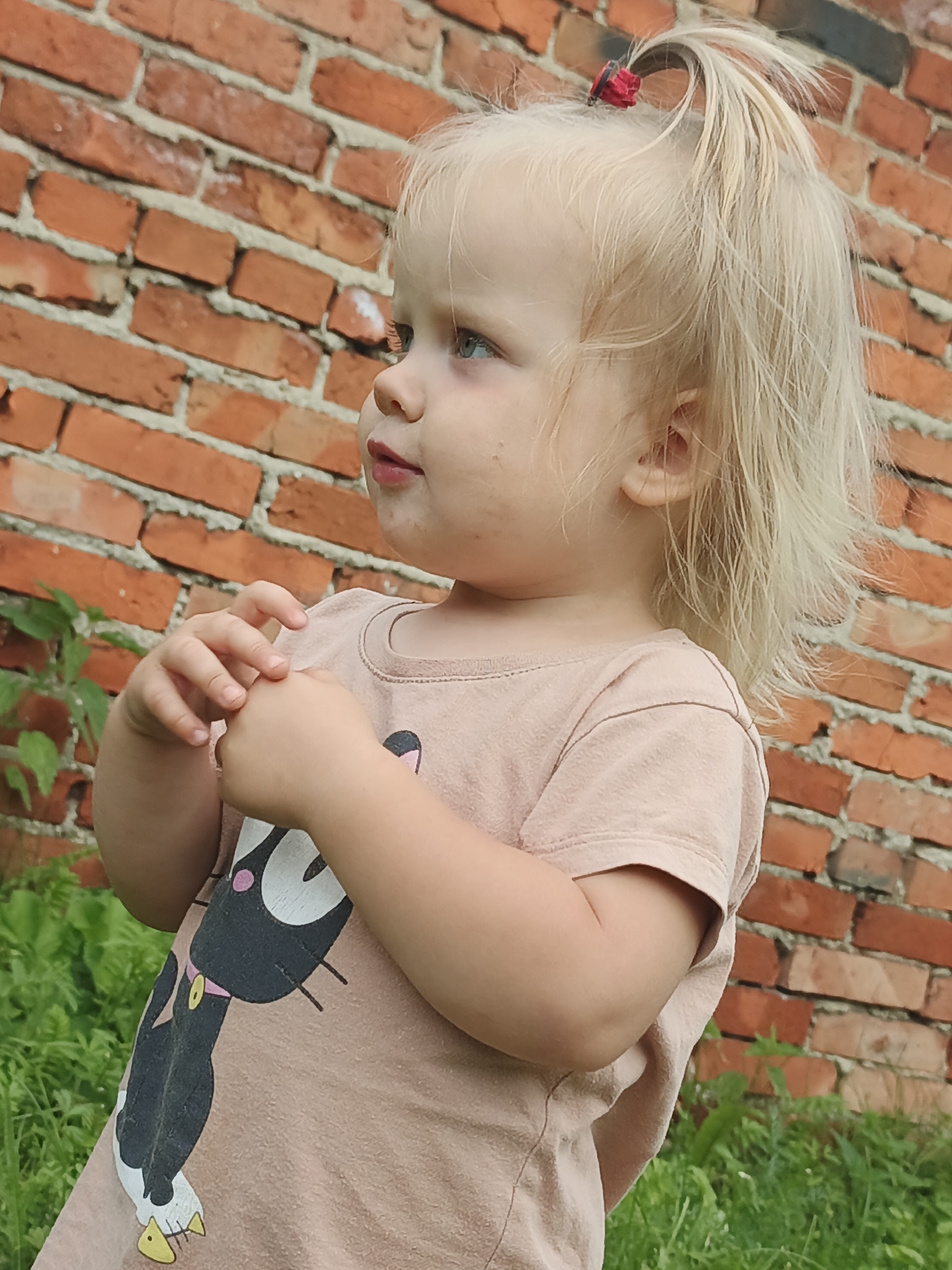 A young Caucasian girl with blonde hair pulled into a ponytail with a red scrunchie stands against a brick wall. She is wearing a light pink t-shirt with a cartoon cat graphic. The girl is looking to her left, with her hands clasped in front of her chest. She is outdoors, with green foliage visible in the foreground and background. The scene is likely in Medvedyovka, Russia.