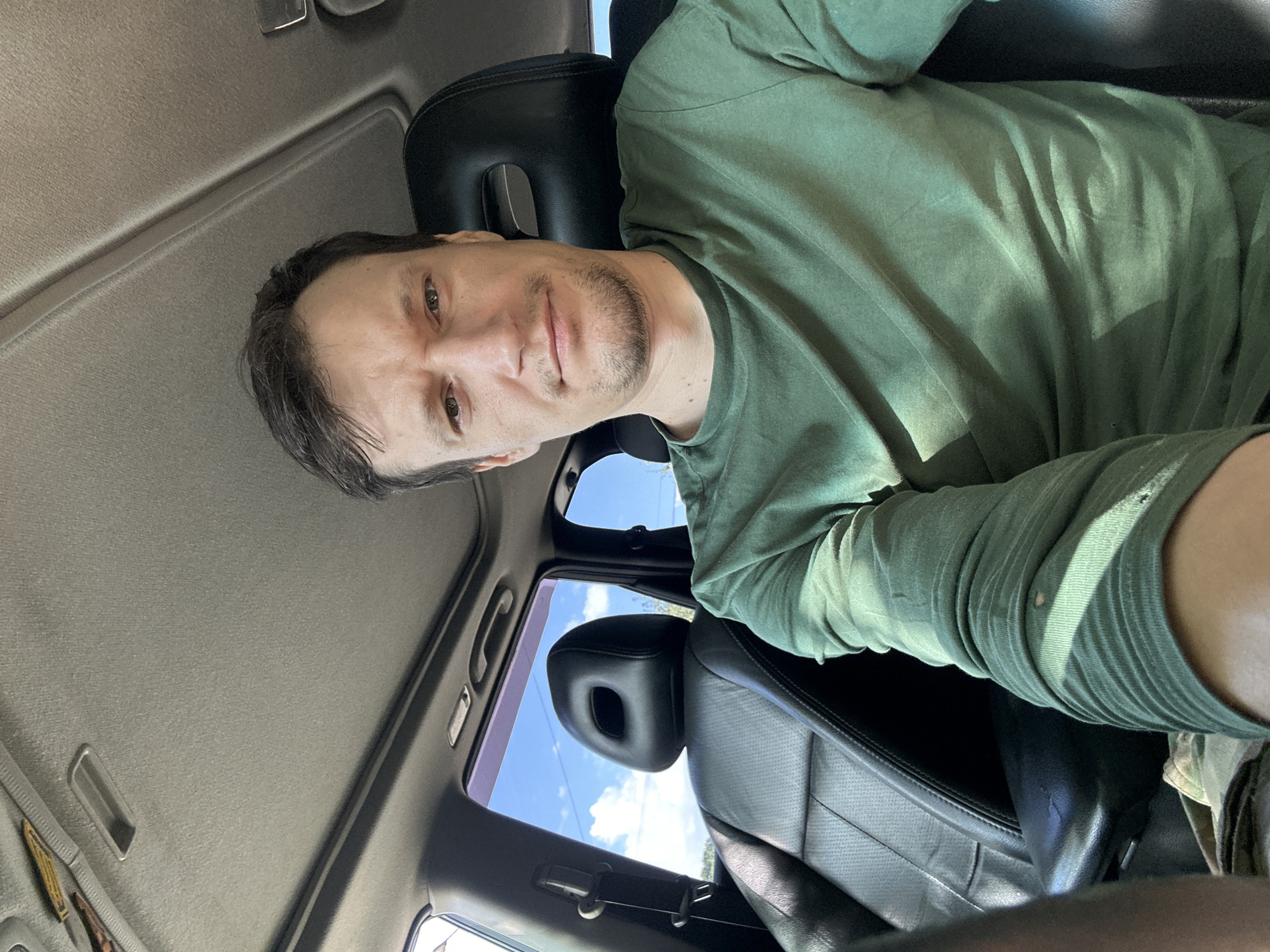 A man with short brown hair and light stubble is sitting in the driver's seat of a car. He is wearing a green long-sleeved t-shirt. The interior of the car is visible, including black leather seats and the sunroof. Outside the car, a blue sky with white clouds is visible through the windows. The image is taken from a selfie perspective, and the man is looking directly at the camera with a neutral expression. There is no indication of the specific location within the city of Makiv, Ukraine, or any forensic details.