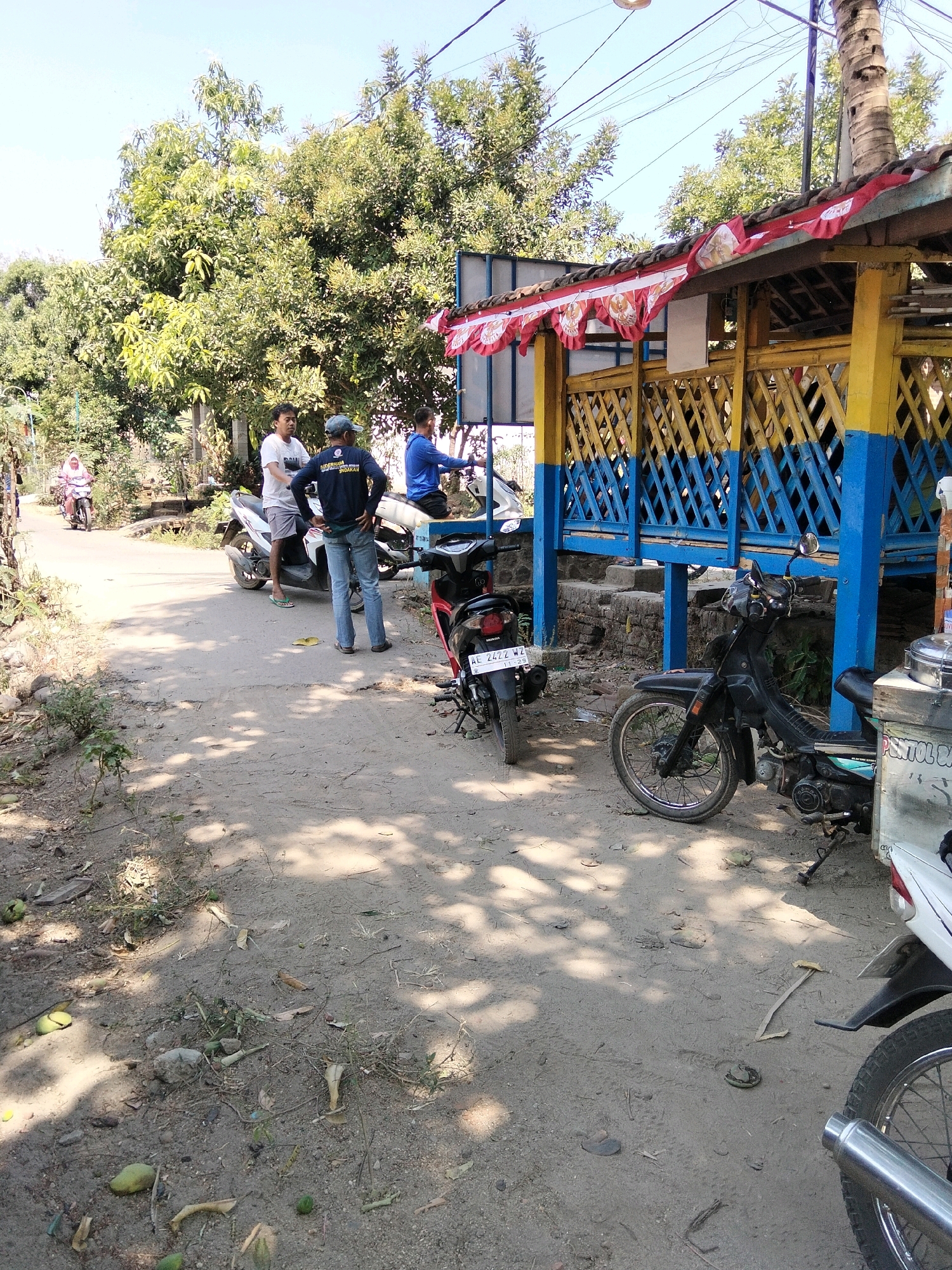 The image depicts a street scene in Jenangan, Indonesia, with a focus on a roadside structure and several motorcycles. In the foreground, a sandy dirt road is visible, with scattered leaves and debris. Two motorcycles are parked on the right side of the road, one black and one white. The black motorcycle is in the foreground, with its front wheel clearly visible. The white motorcycle is partially visible, with its rear end to the right. Behind these motorcycles, a colorful wooden structure with a red and white decorative banner hangs from its roof. The structure features blue and yellow lattice work.
On the left side of the road, three individuals are present. One person, wearing a white t-shirt and shorts, stands near a white motorcycle. Another person, wearing a dark t-shirt and jeans, stands facing away from the camera. A third person, wearing a blue long-sleeved shirt, is seated on a motorcycle further down the road. In the background, a woman wearing a headscarf rides a motorcycle on the road, heading away from the viewer. The environment is characterized by lush green trees, suggesting a somewhat rural or semi-urban setting. The sky is clear and blue.