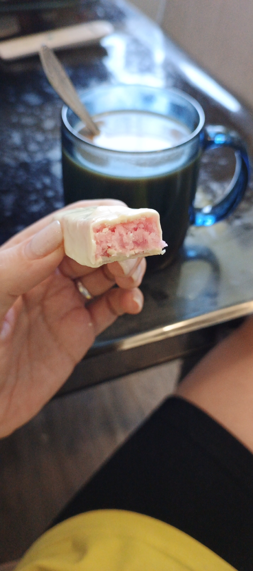 A hand holds a bitten candy bar with a white chocolate coating and a pink, grainy filling. A blue glass mug filled with a dark liquid, presumably coffee, sits on a dark countertop in the background. A spoon rests in the mug. The person is wearing black shorts and a yellow shirt. The location is possibly a kitchen or dining area but the specific location is not identified. The picture does not provide any details to confirm the location in Chaikovsky, Russia.