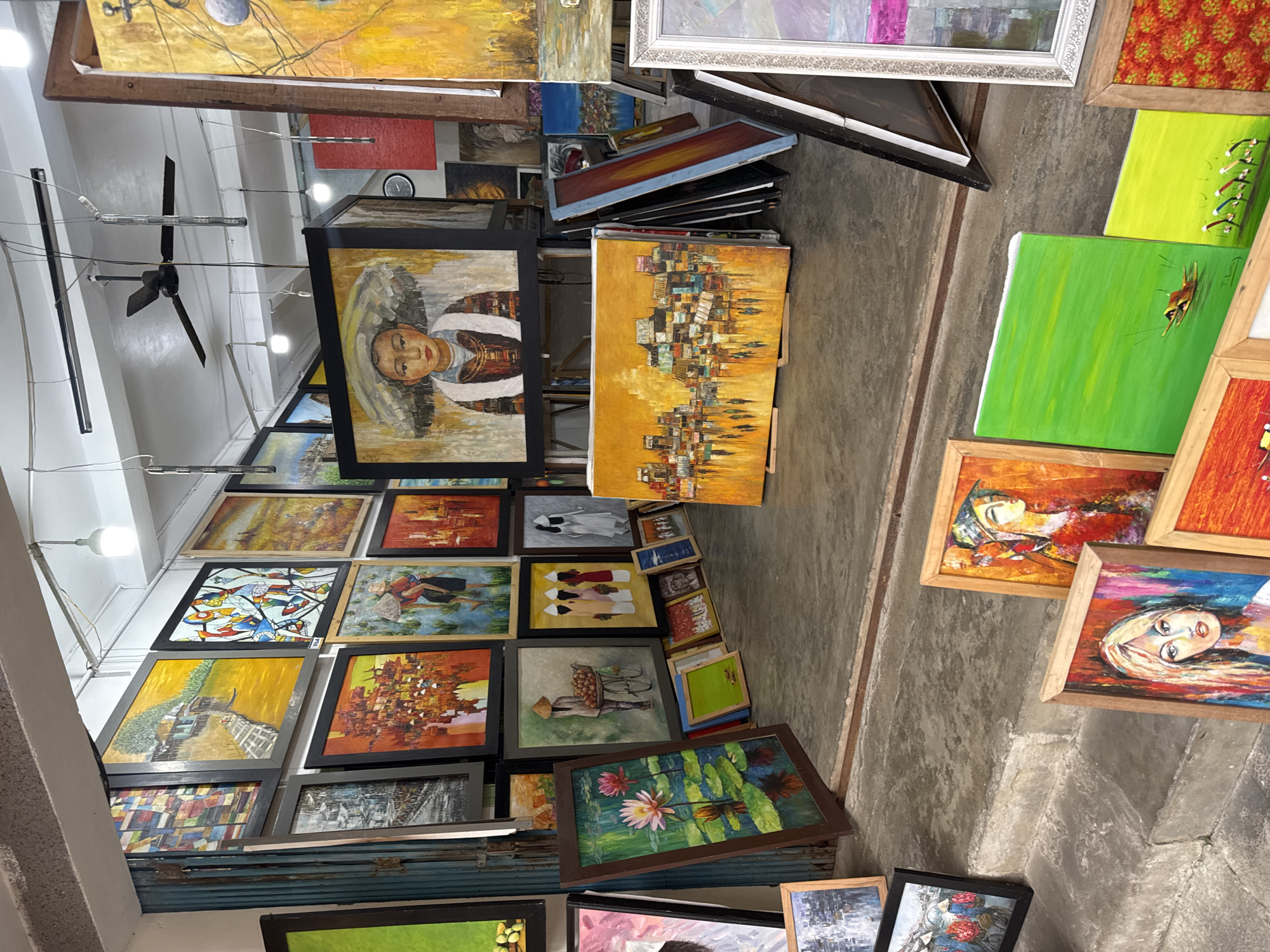 This media file captures an indoor scene, likely an art gallery or shop, filled with a dense display of various paintings. The setting appears to be a practical sales or storage space, with concrete flooring and a white ceiling equipped with fluorescent lighting fixtures and a black ceiling fan.
The dominant subjects are the numerous paintings, which are of diverse sizes, styles, and framed in different materials including black, wooden, and metallic frames. Many paintings are stacked against walls, leaned on the floor, or propped up on makeshift stands, indicating a bustling art retail environment. Visible artworks include:
- Several portraits, such as a prominent one of a person with a serene expression, possibly in traditional attire, framed in black. Another close-up shows a woman's face rendered in vibrant, abstract colors.
- Numerous abstract cityscapes or village scenes, often characterized by warm yellow and orange palettes.
- Nature-themed pieces, such as depictions of lotus flowers on water and green landscapes featuring small boats.
- Figurative scenes showing people engaged in daily activities, like an individual pushing a fruit cart.
- Various abstract artworks with geometric patterns and bright colors.
No specific human activity is captured, but the abundance and arrangement of the artworks suggest a place where paintings are showcased for sale or being prepared for display. A small, round wall clock is faintly visible on a distant wall. The artistic styles, particularly the traditional figures and scenic elements, align with the provided context of Hội An Ward, Vietnam, a region known for its vibrant art scene. No legible text is visible within the image.