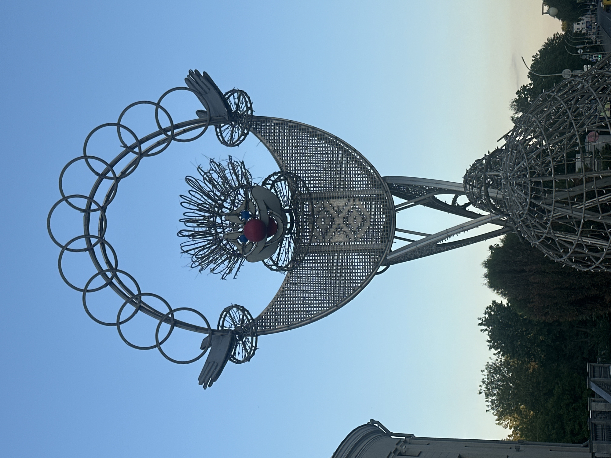 A large, ornate metallic sculpture dominates the frame, set against a clear blue sky. The sculpture is complex, featuring a prominent, upward-curving crescent moon shape crafted from a metal mesh with a grid pattern. Below the crescent, a stylized "head" emerges, characterized by numerous spiky, wire-like protrusions that form hair or a mane, with a distinct red spherical element at its center, resembling a nose or an eye.
An arc of interconnected metal rings, similar to the Olympic symbol, extends from and encircles this "head," acting as a halo or crown. Two flat, stylized hand-like forms are attached to the outer ends of this ring structure. The entire upper portion of the sculpture is supported by a tall, tapering base or column, also constructed from interconnected metal elements. To the right of the main structure, a large, partially visible spherical or ovoid metal framework, made of numerous interconnected bars, suggests it might be an adjacent component of the same installation.
The background transitions from a deep blue at the top to a lighter hue closer to the horizon. Lush green trees and foliage are visible on the right side and in the lower background, indicating a park-like setting. Faint outlines of urban infrastructure, including buildings and possibly streetlights, can be seen in the distant background. The soft, even lighting and cloudless sky suggest either early morning or late afternoon. No people or visible text are present. This sculpture is located in Minsk, Belarus, and is commonly known as "Grandfather Arkhip."
