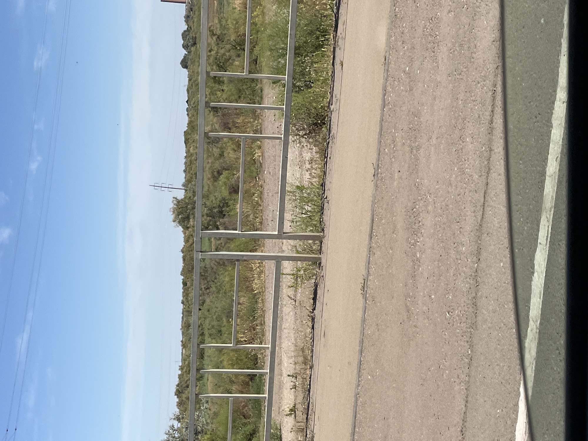 A daytime exterior shot features a metal fence in the foreground, separating a paved area from a vegetated landscape. The fence is made of silver-colored metal bars arranged in a rectangular grid pattern. Behind the fence, the landscape consists of dry grass and bushes, with some taller trees in the background. The sky is clear blue with a few wispy clouds. In the distance, a tall, reddish-brown industrial chimney is visible on the right side of the image. Power lines stretch across the sky, and a utility pole stands to the left of the chimney. The paved area in the foreground appears to be a road or pathway, with a white line marking its edge. The overall scene suggests an outdoor environment, possibly on the outskirts of an urban or industrial area.