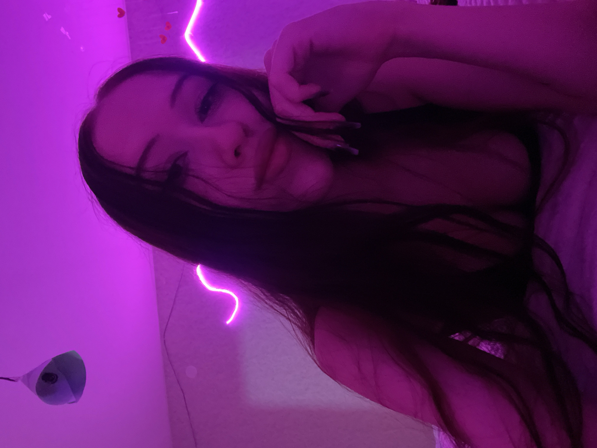 The image shows a close-up of a young woman's face and upper body, illuminated by pink and purple lighting. She has long, dark hair and is resting her chin on her hand. The background includes a textured wall with some decorative pink neon lights and small heart decorations. A light fixture is visible hanging from the ceiling in the upper left corner. The overall impression is that of a selfie taken in a bedroom setting. There is no visible indication of the specific location within Votkinsk, Russia, or any details about the woman's actions or interactions.