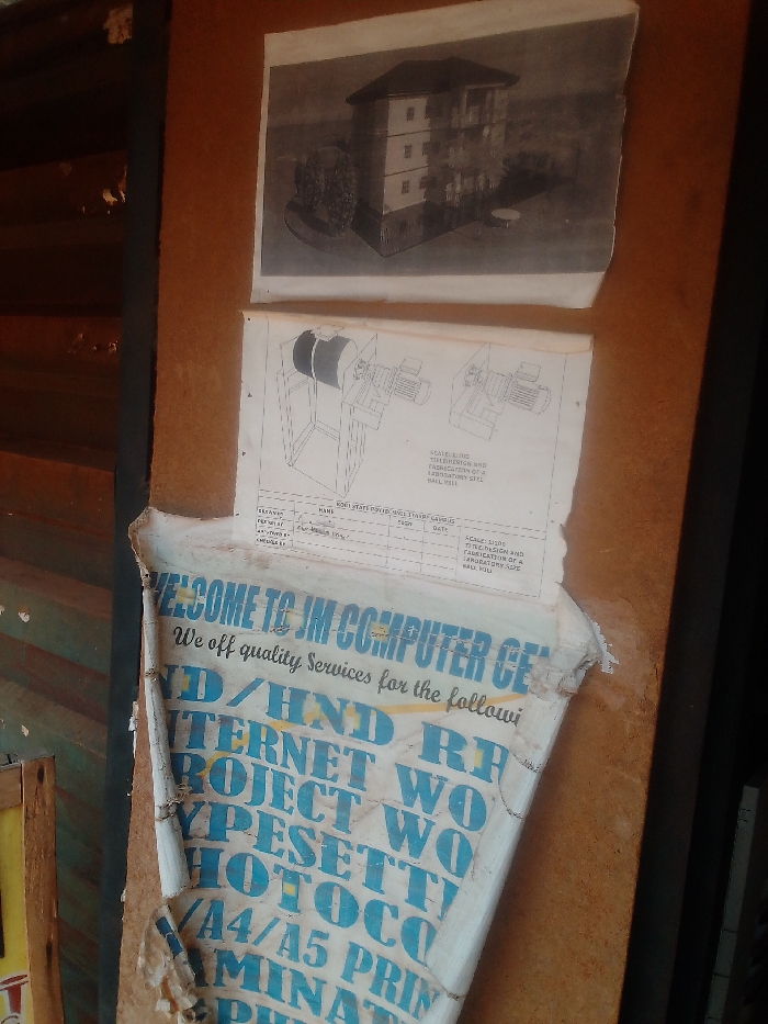 The image depicts a wooden surface with three pieces of paper pinned to it. The topmost paper is a black and white photograph of a three-story building with a green bush in front of it. The middle paper is a diagram of a piece of machinery with text that reads "Scale: 1:100, Design, Implementation and Fabrication of a Laboratory Size Ball Mill." The bottom paper is a torn, faded, and yellowed advertisement for a business called "JM Computer Center" in blue and yellow lettering. The advertisement advertises services including "D/HD," "Internet RF," "Project Work," "Typeset Work," "Hot Occ," "A4/A5 Print," and "Laminate." The location is Auchi, Nigeria, based on coordinates (latitude: 7.062, longitude: 6.273).