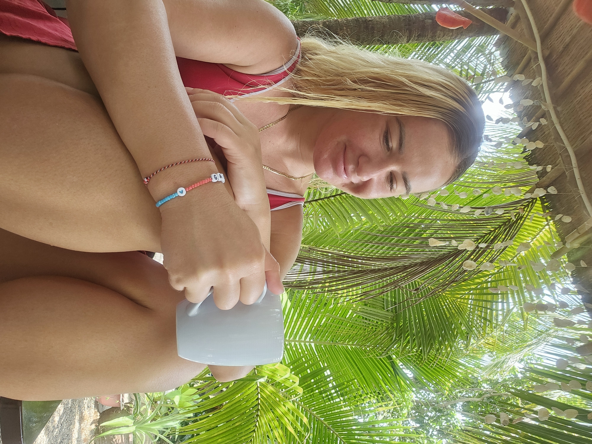 In the city of Phu Thuy Ward, Vietnam, a young woman with blonde hair sits under a thatched roof adorned with shell decorations. She is wearing a red swimsuit and has a light pink facial mask on. The woman is holding a white mug in her hands, which are adorned with bracelets. She is looking down and smiling, seemingly relaxed. The background is filled with lush green palm leaves, suggesting a tropical outdoor setting. The scene is captured from a medium shot, focusing on the woman and her immediate surroundings.