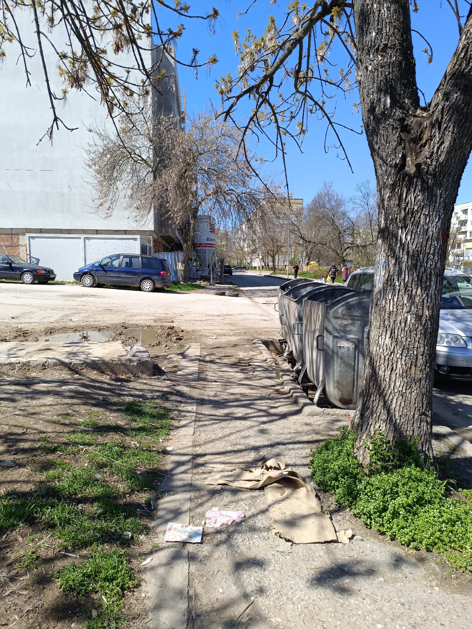 The image depicts an urban scene in Pleven, Bulgaria. The environment is somewhat unkempt, with a damaged road, puddles, and litter visible. The scene is characterized by a mix of natural and industrial elements.
**Location:** Pleven, Bulgaria
**Objects:**
* Several cars, including a dark-colored sedan and a blue minivan, are parked along the street. A silver car is also parked next to trash containers.
* Large, metal trash containers are lined up along the sidewalk.
* A large tree dominates the foreground on the right, casting shadows. Another tree is visible towards the background.
* A building with a tall, cylindrical structure extending upwards is visible.
* A billboard with Cyrillic text is partially visible.
* Debris, including pieces of cardboard, paper, and other litter, scatters the ground.
**People:**
* A small group of people are walking along the sidewalk.
**Environment:**
* The weather appears to be clear and sunny, based on the shadows and blue sky.
* The overall impression is one of neglect and disrepair.
* Potholes with pooled water are visible.
* Vegetation, including grass and small plants, is present.