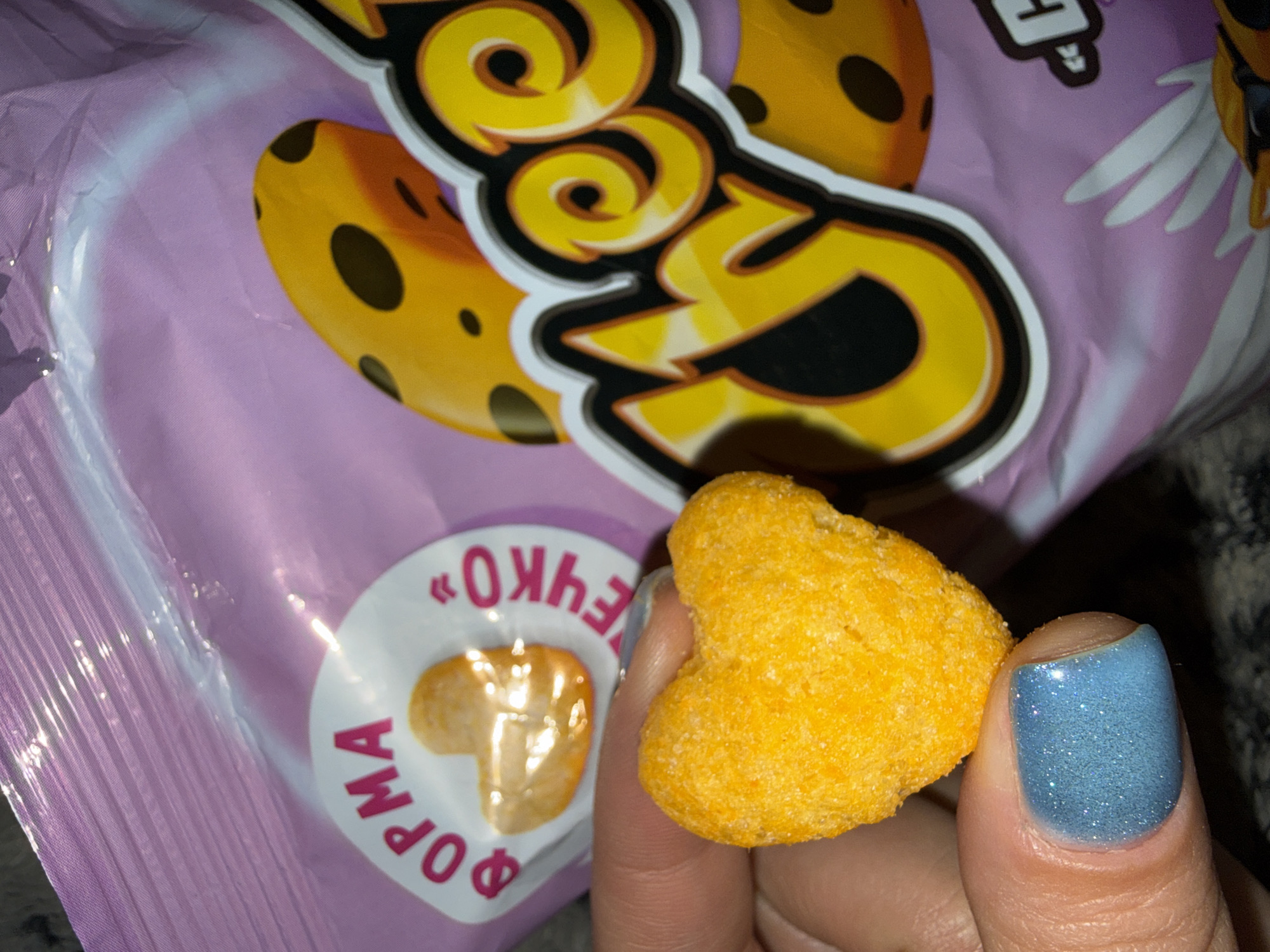 A close-up photograph captures a hand holding a heart-shaped, bright orange, puffed snack against the backdrop of a crinkled purple snack bag. The snack is visibly coated with a fine, powdery seasoning, characteristic of cheese-flavored snacks. The person's thumb, adorned with shimmering light blue nail polish, securely holds the snack.
The snack bag, made of a glossy, reflective plastic material, displays prominent branding. A large, stylized yellow logo with white and black outlines dominates the upper portion of the bag, strongly resembling the "Cheetos" brand logo, possibly in Cyrillic characters with distinctive swirls and curls. To the right of this main logo, a cartoon character with wings and sunglasses is partially visible, also typical of Cheetos branding.
On the lower left of the bag, a white circular sticker is prominently featured. This sticker contains Cyrillic text: "ФОРМА" (FORM, or SHAPE) is visible along its bottom curve, and "ОЧЕНЬ" (VERY) is visible along its top curve, likely emphasizing the snack's unique shape. Inside the circle, a smaller, heart-shaped snack, identical in appearance to the one being held, is depicted.
The setting appears to be indoors, under artificial lighting, which creates reflections on the plastic bag and the person's glossy nail polish. The image captures a casual moment of enjoying a snack, likely in Dmitrov, Russia, given the provided context.