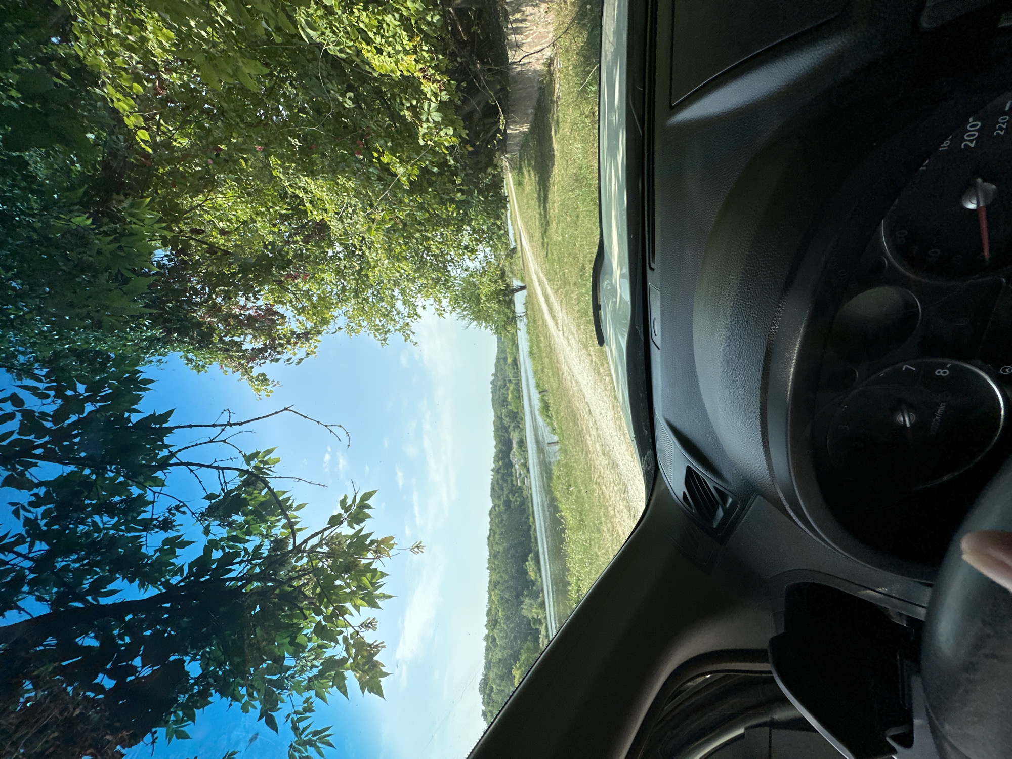 This is a first-person perspective shot from the inside of a vehicle, looking out through the windshield. The driver's side dashboard and steering wheel are visible in the foreground, with a finger in the lower left corner. The vehicle appears to be on a dirt road next to a body of water, possibly a river or lake. Lush green trees and vegetation line the right side of the road, with some red berries visible on the branches. The sky is a bright blue with some scattered clouds. In the distance, a tree-covered hillside can be seen across the water. The scene suggests a rural or natural setting, possibly in the city of Makiv, Ukraine, given the context.