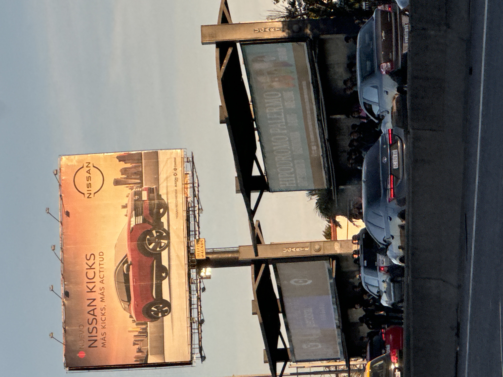 The image captures a roadside scene in Olivos, Argentina, featuring several prominent elements. There is a large billboard advertising the "Nissan Kicks," another advertising "Hipódromo Palermo" billboards with "Ugarte" signs on top, and traffic on a road adjacent to the signs. The lighting suggests late afternoon, and the sky is clear. There are several cars, and also what appear to be people walking or standing nearby, potentially in a line or crowd.