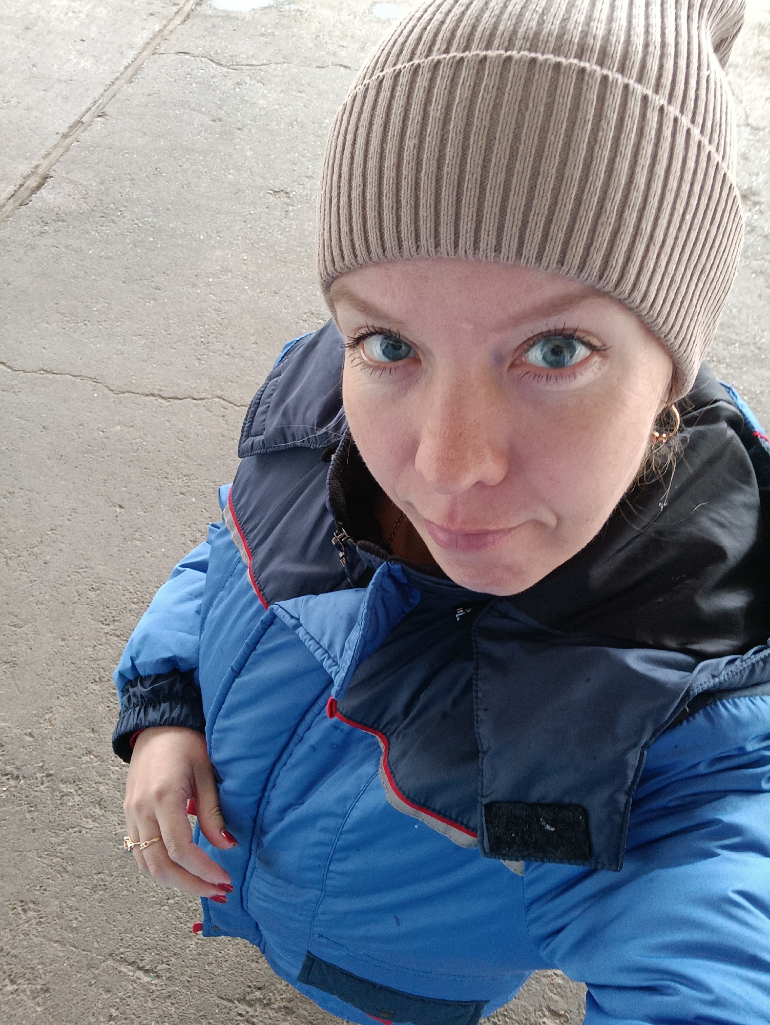 A person wearing a beige ribbed beanie and a blue puffer jacket with a dark blue collar stands on a grey concrete surface. The person has fair skin, blue eyes, and red nail polish. A gold ring is visible on one of their fingers. The image is a high-angle shot, with the person looking up at the camera. The location is consistent with an outdoor area with concrete surfaces. Based on the request for location details, it may be in the city of Chaikovsky, Russia, but no specific identifiers confirm this.