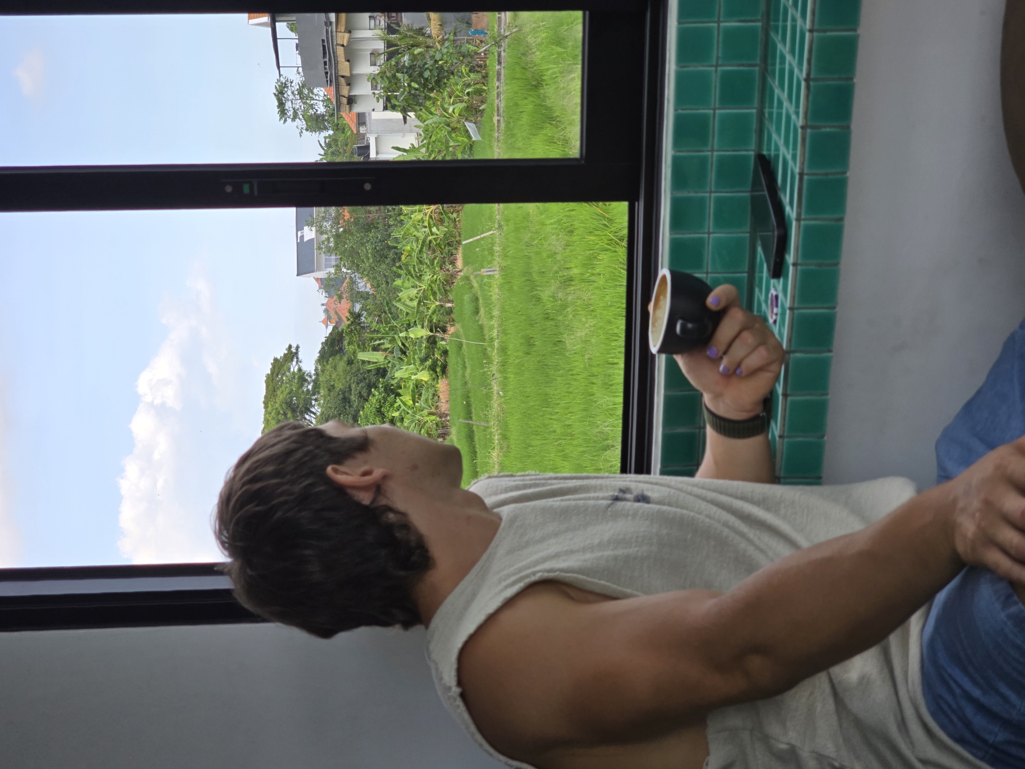 A sideways view reveals a man, likely Caucasian, with brown hair and a fair complexion, sitting near a window in what seems to be an indoor space. He is wearing a light-colored, sleeveless shirt and blue pants. He's holding a small, black coffee cup in his left hand, with his hand showing painted fingernails. He's looking out the window. A black wrist watch is visible on his left wrist.
The window frames a view of a vibrant green landscape, possibly a rice field, with trees and building structures in the background. The tiled wall next to the window features green square tiles.
Based on the presence of rice fields and a combination of rural and urban elements, this image could be taken in North Kuta, Indonesia. A phone is positioned along the wall with green tiles.