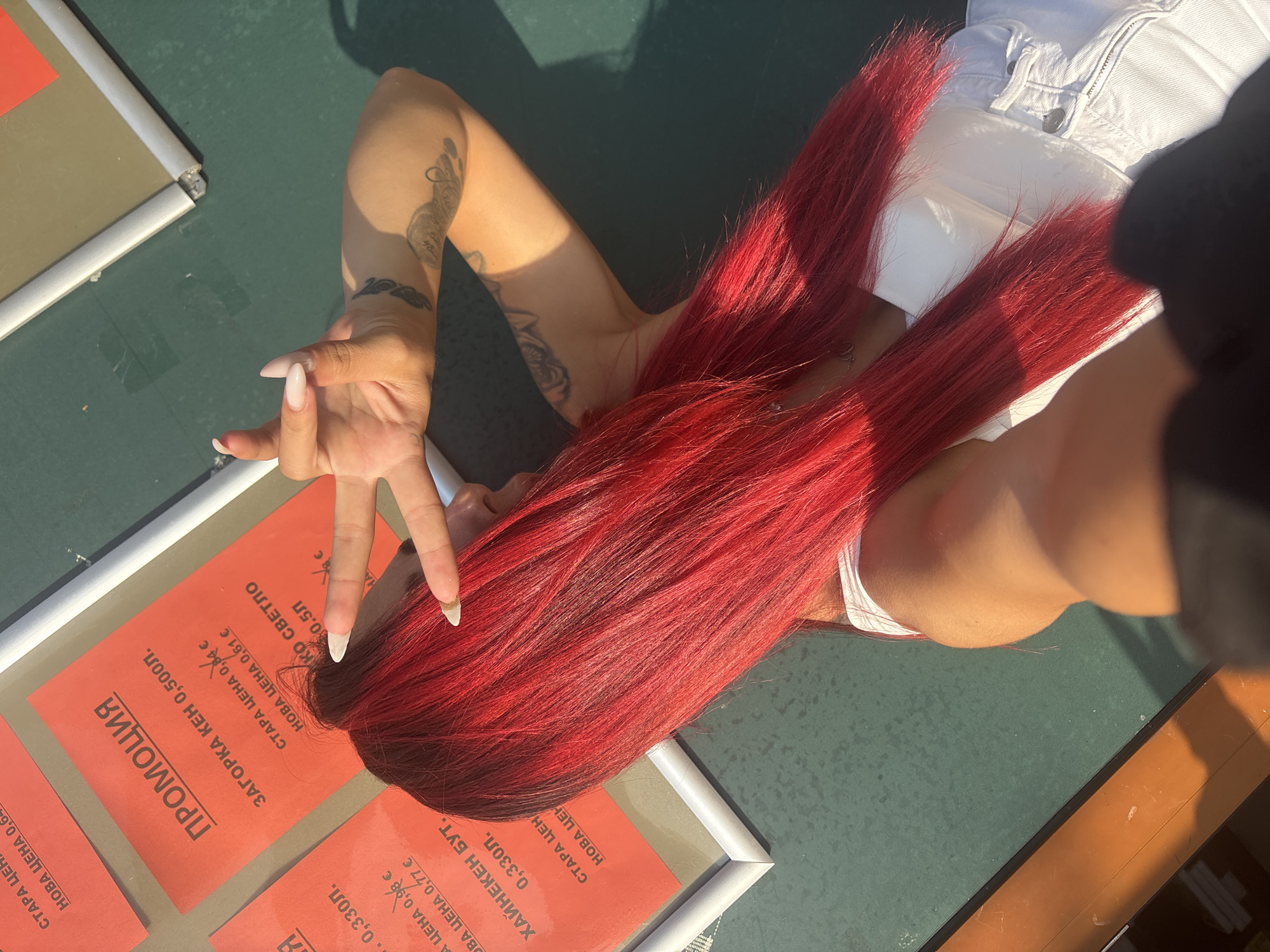 In Sofia, Bulgaria, a young woman with vibrant red hair is posing for a photo in an outdoor setting. She is wearing a white top and white jeans. Her right hand is raised, making a peace sign. Behind her, several red promotional flyers are visible, attached to a green surface. One flyer prominently displays the word "ПРОМОЦИЯ" (Promotion) in large black letters, followed by details about product prices in Bulgarian currency, such as "ЗАГОРКА КЕН 0,500л." and "СТАРА ЦЕНА 0,84 €." Another flyer mentions "ХАЙНЕКЕН БУТ. 0,330Л." and "СТАРА ЦЕНА 0,98 €." The scene is illuminated by sunlight, casting shadows. The overall impression is a casual, candid shot capturing a moment of leisure or shopping.