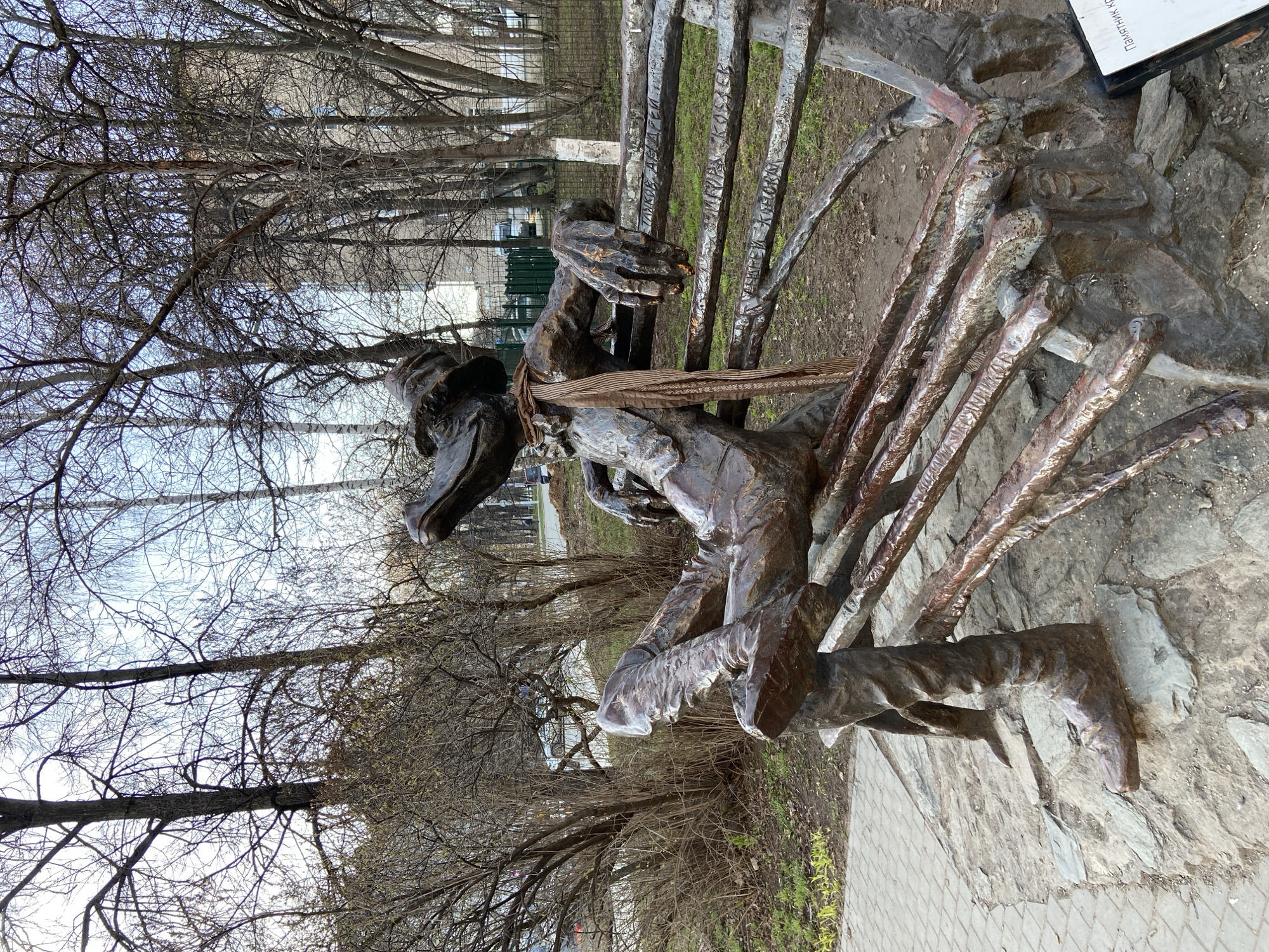 A bronze-colored metal sculpture depicts a seated figure on a park bench. The figure, dressed in a hat, has a real brown and beige striped fabric scarf tied around its neck. Its hands are clasped together, resting on its lap. The bench, crafted from the same dark metal, has horizontal slats forming its backrest. Inscriptions are visible on two of these slats: "ДАЛИЯ АБДУЛЛАЕВ" and "ИЛИМЕРА КАСАЕВ".
The sculpture is positioned outdoors on a base of irregularly shaped flagstones and dirt, adjacent to a paved walkway made of interlocking pavers. The background features numerous bare deciduous trees and shrubs, indicative of late winter or early spring, under a bright, overcast sky. Patches of sparse, dry grass are visible on the ground. A small, partially visible white information plaque rests near the sculpture's base, with the text "ТОМЕТИК-К..." somewhat discernible. The scene is illuminated by daylight, creating a quiet, contemplative atmosphere in Izhevsk, Russia.