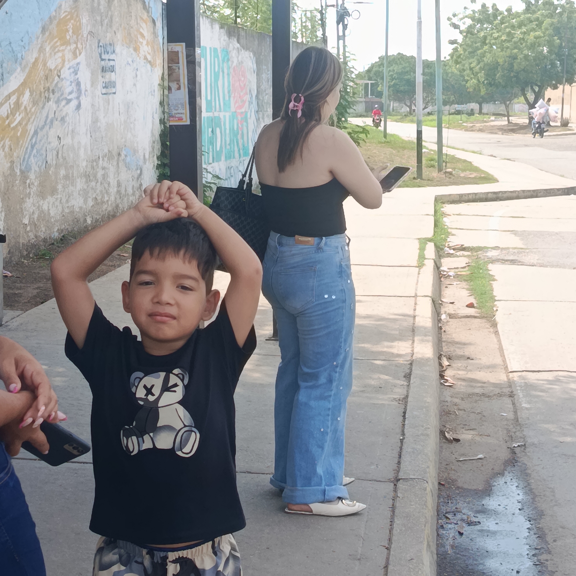 A medium outdoor shot shows a child and an adult standing on a sidewalk next to a street. The child, a boy with dark hair, is in the foreground, wearing a black t-shirt with a cartoon bear graphic and patterned shorts. He has his hands raised and clasped above his head. Behind him is an adult woman with long brown hair pulled back with a pink clip. She is wearing a strapless black top, light blue jeans, and white shoes. She is holding a cell phone. To the left, a partial view of another woman’s hand holding a cell phone is visible. A black purse hangs on the woman's shoulder. The sidewalk is next to a street with two people on motorcycles in the distance. A concrete wall with peeling paint is behind the woman. The location is identified as Los Guayos, Venezuela, based on contextual clues like architectural styles and typical environmental characteristics.