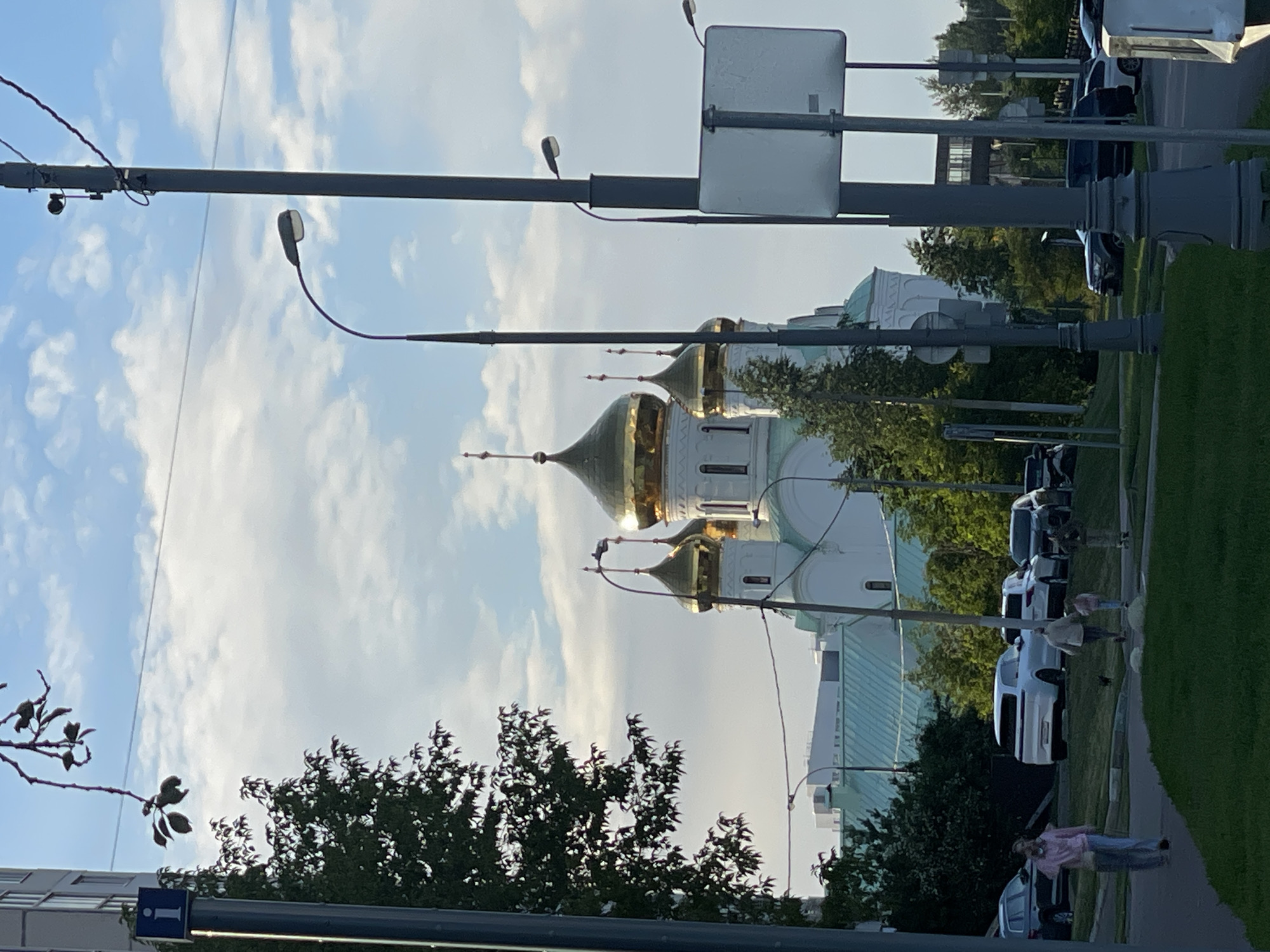 This image depicts an outdoor scene in Moscow, Russia, likely during the daytime. The focus is on a large white building with distinctive golden domes, characteristic of Orthodox churches. The building's architecture is intricate, with multiple dome structures visible, some topped with crosses.
In the foreground, several streetlights and utility poles are present, some with wires strung between them. There are also several cars parked along what appears to be a street or parking lot adjacent to the building and a grassy area. Some trees are visible, providing a natural element to the urban landscape.
Several people are present in the scene. One person is walking on a paved path, while others appear to be near parked cars. The people's activities are not clearly defined, but they seem to be engaged in everyday outdoor pursuits. The lighting suggests it is daytime, possibly late afternoon or early evening due to the soft light and shadows. The sky is partly cloudy, indicating fair weather.
A blue sign with a white "i" symbol is visible on one of the poles, which typically indicates an information point. The overall setting is a mix of religious architecture, urban infrastructure, and natural elements, common in many Russian cities. No specific event or emotional context is evident beyond a general sense of a normal day.