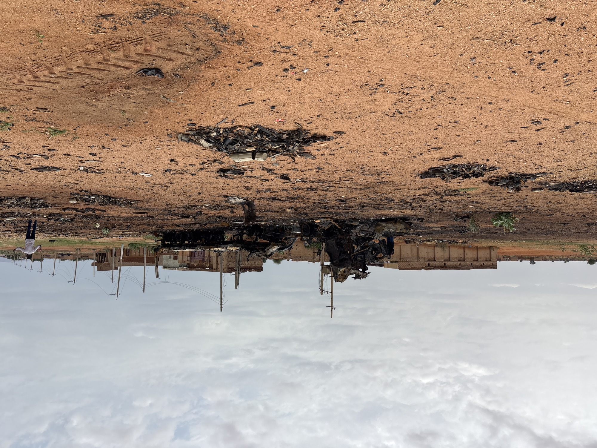 In Um Laba, Sudan, the scene depicts the aftermath of a significant incident, with two individuals present. One person, dressed in dark clothing, stands near a heavily damaged vehicle in the mid-ground. Further to the right, another individual in a white shirt and dark pants gestures with their arms outstretched. The environment is an open, arid area with red soil, scattered debris, and remnants of burnt structures in the background. The foreground is littered with ash, burnt material, and tire tracks, suggesting recent destruction. A partially intact building with a long, low facade is visible behind the primary wreckage, along with utility poles and power lines extending into the distance. The sky is overcast with clouds.