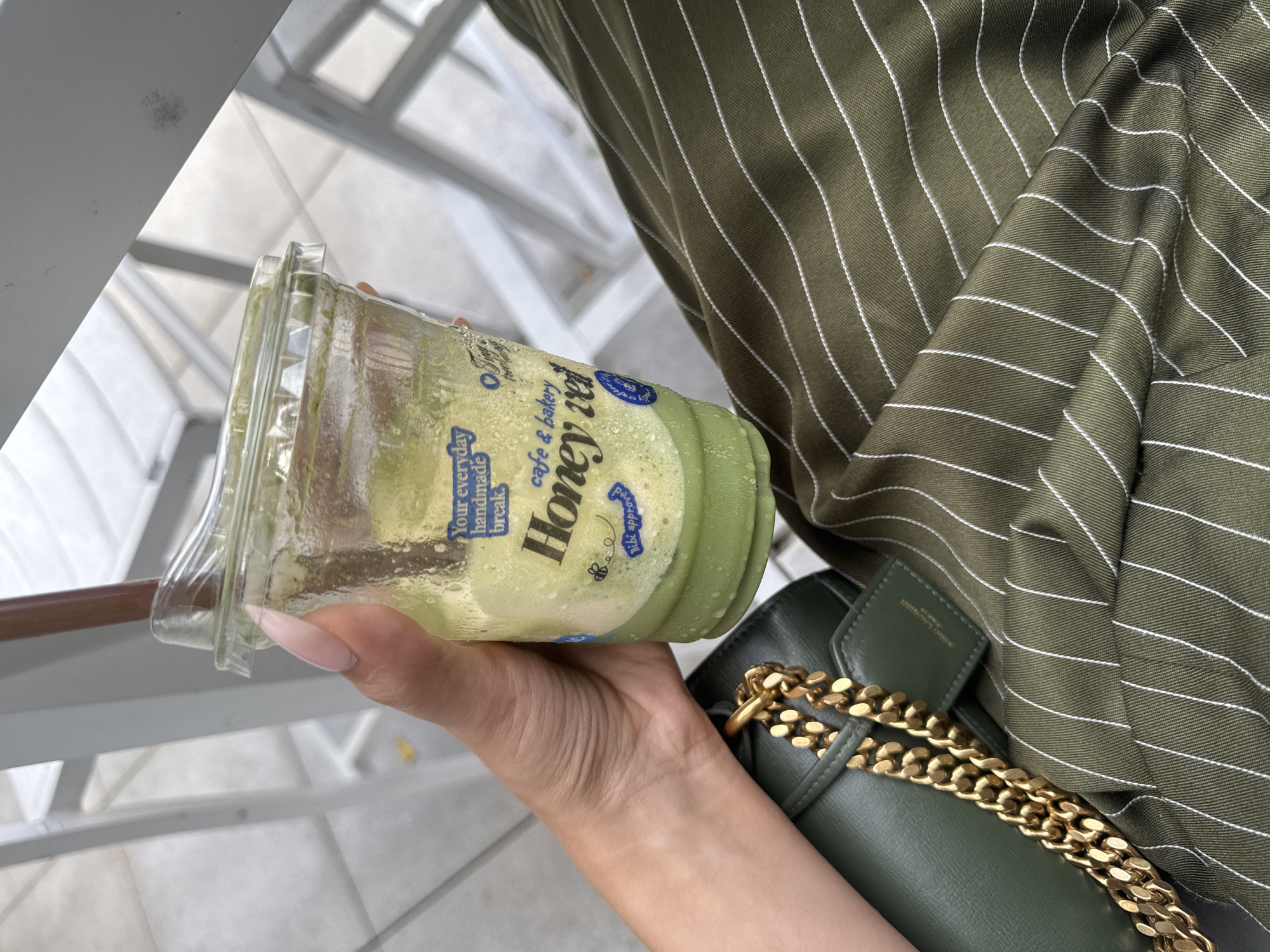 A person's hand with light-colored, manicured nails holds a clear plastic cup filled with a green, creamy beverage. Condensation beads on the outside of the cup. The cup has a blue label with white text that reads "Your everyday handmade break" and "Honey rev cafe & bakery". Below "Honey rev", there is a small drawing of a bee. The person is wearing a dark green, pinstriped shirt. A dark green purse with a thick gold chain strap is visible next to the cup and shirt. The background appears to be an indoor or outdoor setting with bright, natural light, suggesting daytime. The metallic railings and tiled flooring visible in the background suggest a public area like a mall, cafe, or beachside promenade. The overall mood is casual and relaxed, perhaps a moment of enjoying a refreshment. The text indicates the beverage is from "Honey rev cafe & bakery". No specific time of day or weather conditions are explicitly mentioned beyond the bright lighting.