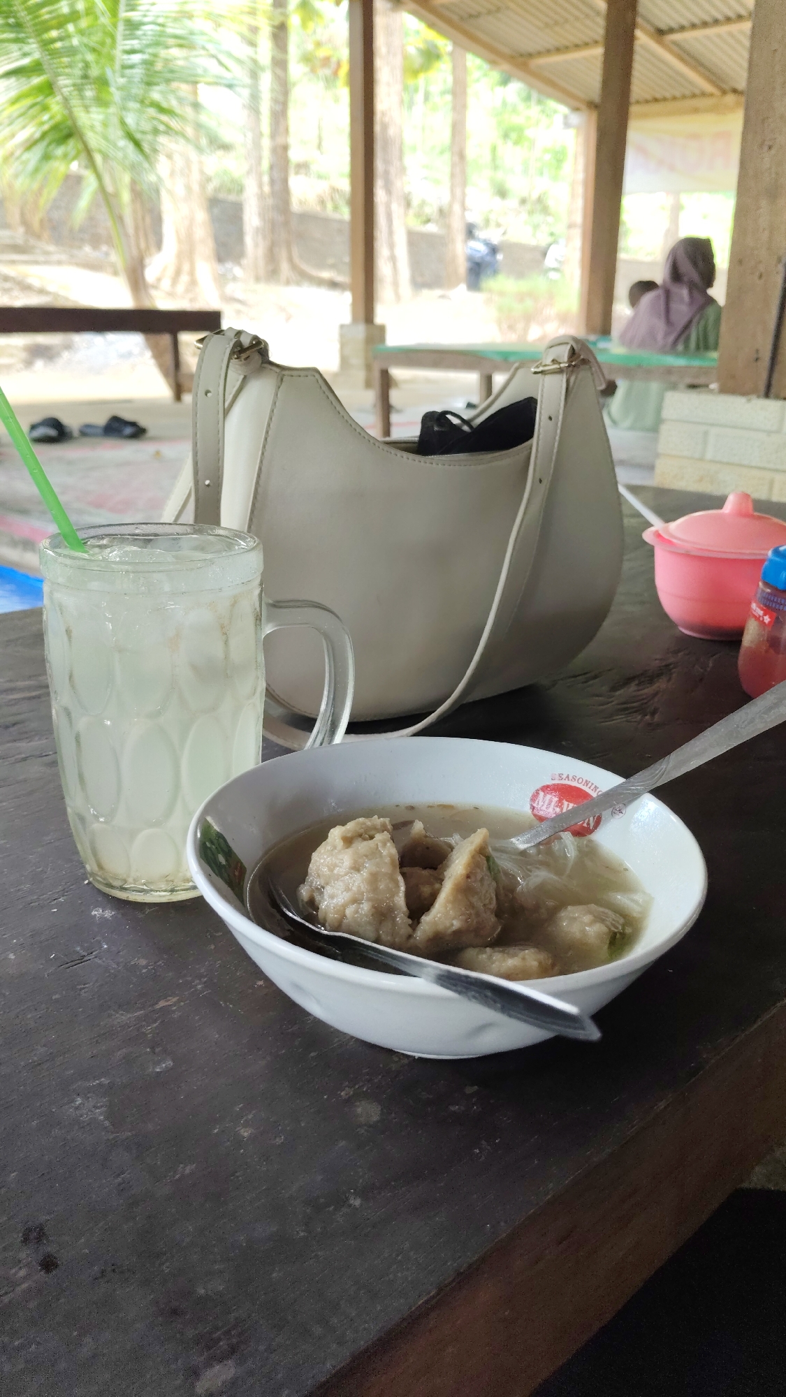 An overhead medium shot captures a wooden table on the veranda of a restaurant in Kresek, Indonesia, with a glass of iced beverage, a bowl of meatballs, and a beige handbag as the primary subjects. The glass, positioned to the left, is filled with a light-colored beverage and ice cubes, with a green straw protruding from the top. Adjacent to it, on the right, is a white bowl containing several meatballs in broth, with a silver spoon resting inside. A beige handbag is placed behind the food and drink. In the background, out of focus, is a person wearing a headscarf and a green garment, seated at a table. The environment appears to be an open-air dining area, with wooden pillars and a corrugated metal roof visible. The table itself is dark wood, showing signs of wear and tear.