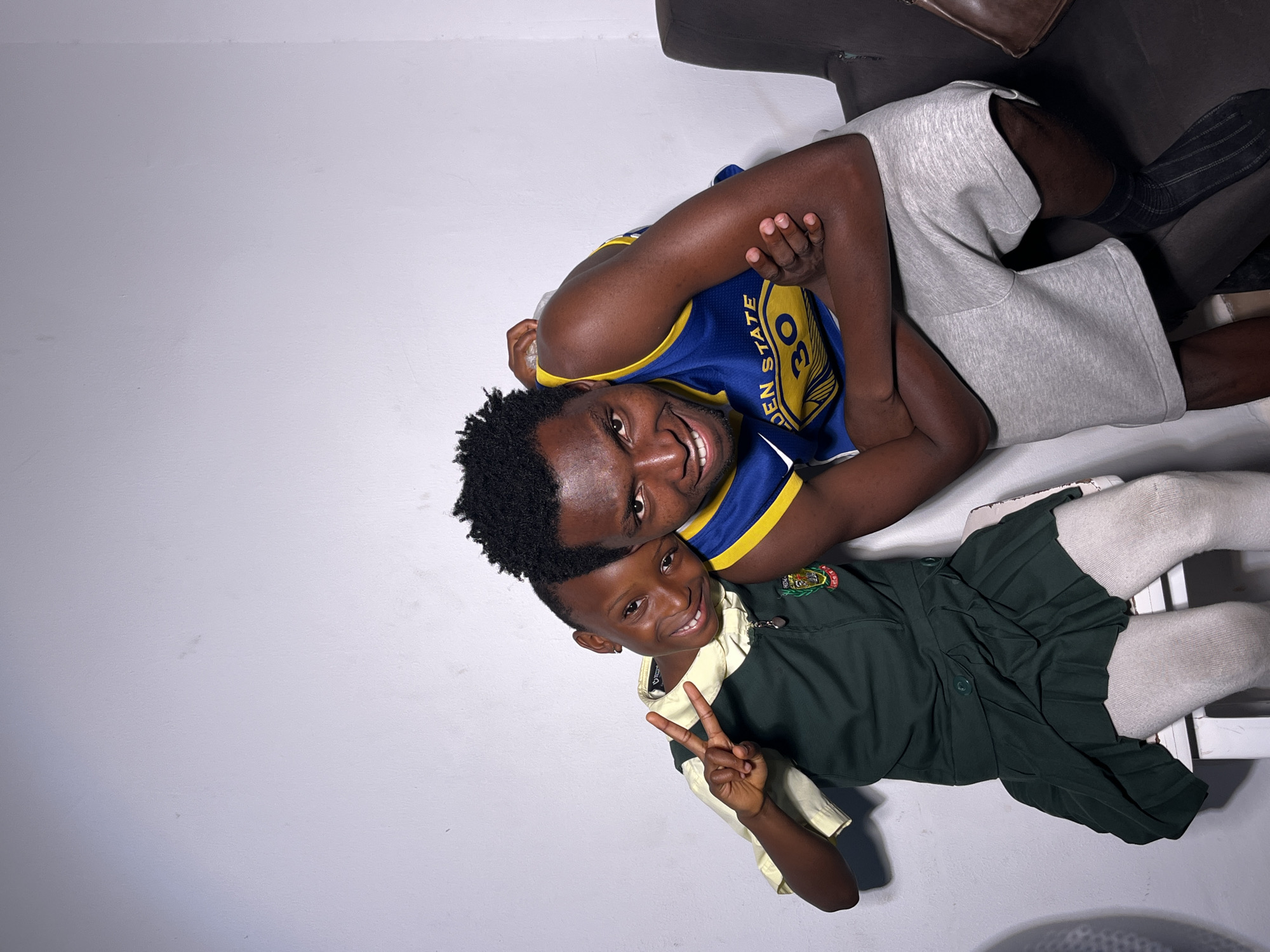 Two people, an adult and a child, are posing together. The adult is wearing a blue and yellow Golden State Warriors jersey with the number 30. They are also wearing light gray shorts and dark socks. The child is wearing a dark green school uniform with a light yellow shirt underneath, a pleated skirt, and white socks. The child is making a peace sign with their fingers. Both individuals appear to be smiling, suggesting a happy or playful mood. They are positioned on a plain, light-colored surface, possibly a floor or a bed, with a dark piece of furniture in the background. The lighting is bright, casting soft shadows. There is no discernible indication of the time of day, weather, or specific location beyond the indoor setting. The image has a casual and intimate feel, possibly depicting a moment between family members or friends.