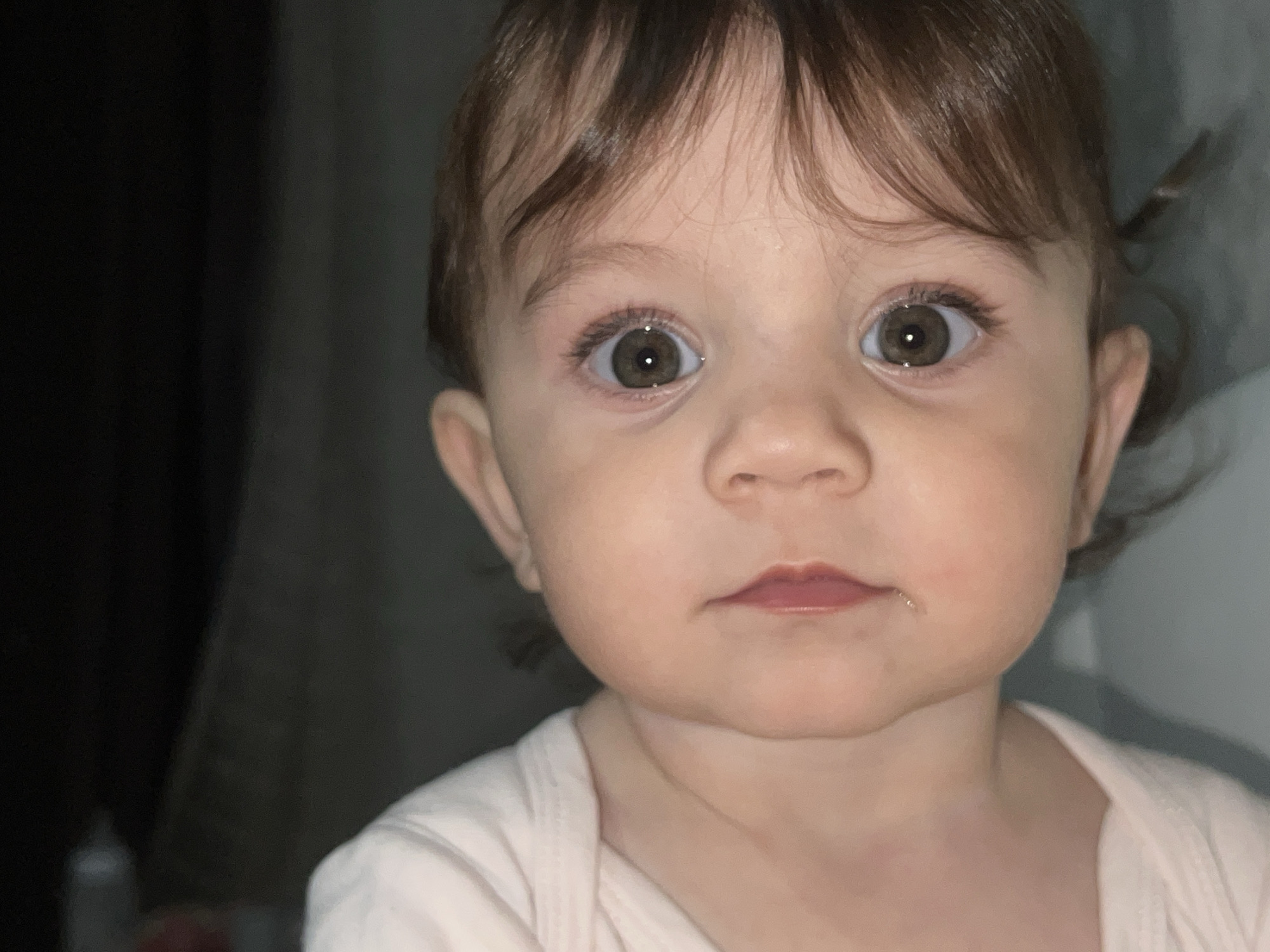 A close-up portrait of a young child with brown hair and wide, brown eyes. The child has fair skin and a small, slightly parted mouth. The child is wearing a light-colored shirt. The background is dimly lit, with a dark area on the left and a lighter area on the right. There is no information to identify the location as Elena, Bulgaria.