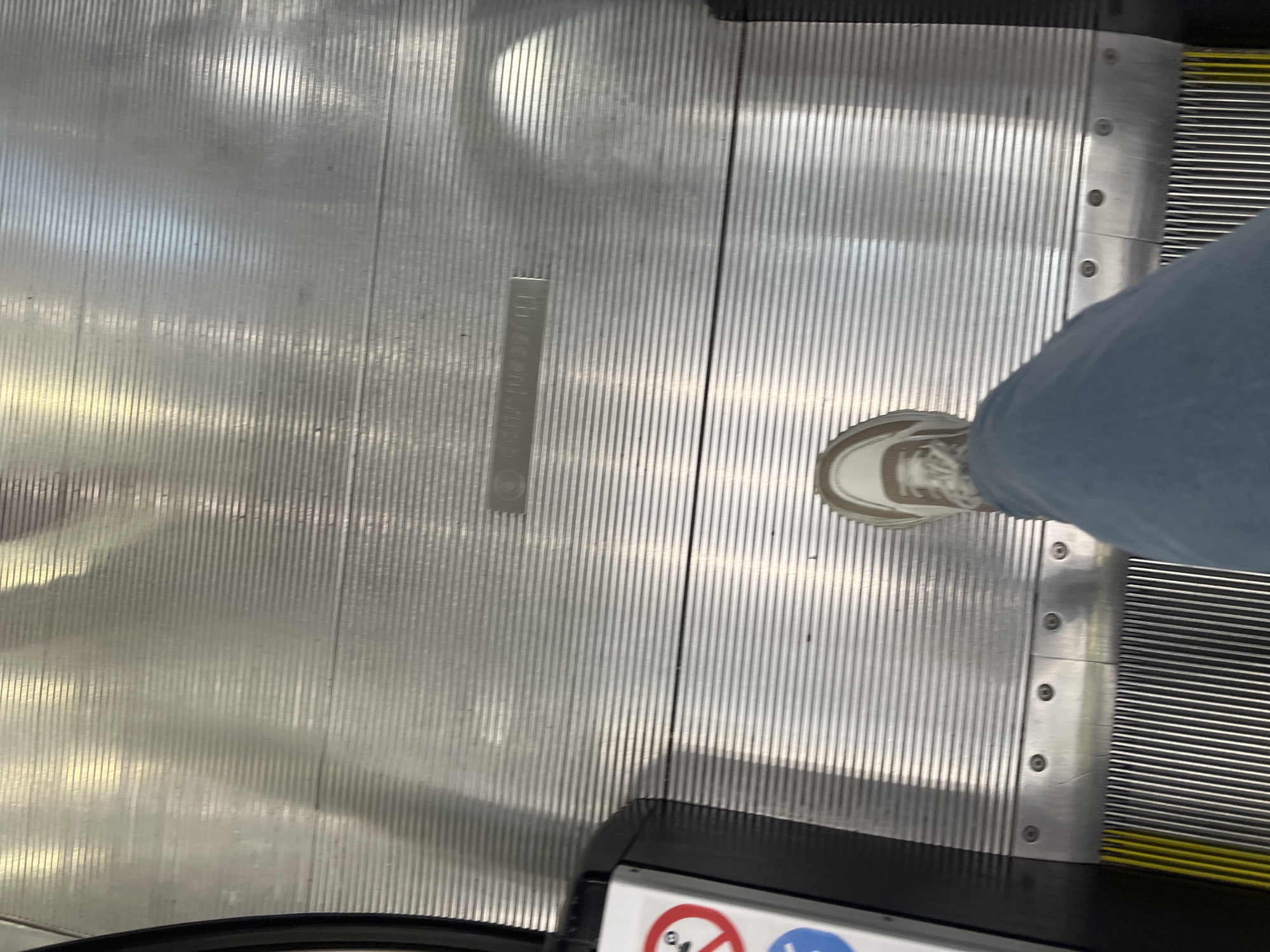 The image shows a first-person perspective of someone standing on an escalator. The escalator steps are made of metal with horizontal ridges. The person is wearing blue jeans and a white and brown sneaker. To the left, there is the black side of the escalator with a white panel that has red and blue warning symbols. The scene is indoors, likely in a public place such as a shopping mall or airport. There is no identifiable information to determine the specific location within Yekaterinburg, Russia.
