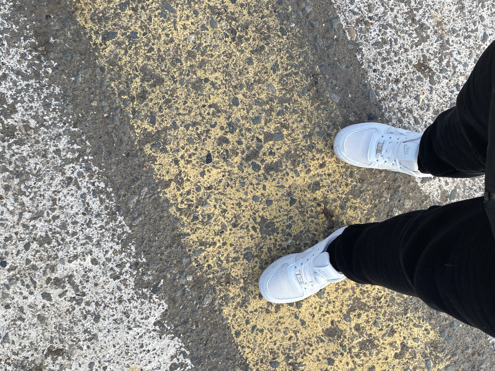 An overhead shot captures the lower legs and feet of a person standing on an asphalt surface. The individual is wearing black trousers and white sneakers. The right sneaker clearly displays the "NIKE" logo on its tongue. The ground features worn, textured asphalt, primarily dark grey with visible small stones. Painted markings are prominent on the ground: a wide, faded yellow stripe extends horizontally across the middle of the frame, underneath and around the person's feet. To the left and right of this yellow section, the asphalt is covered with irregular patches of faded white paint. The overall impression is of worn road or parking lot markings. The lighting is bright and even, suggesting it is daytime. No specific activity beyond standing or walking is discernible. The scene is set in an outdoor urban or semi-urban environment in Марковский, Russia.