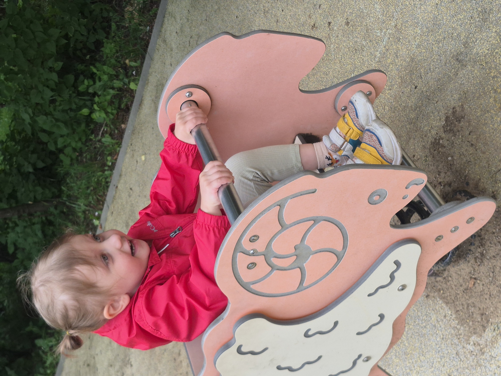A young child with light brown hair styled in a small ponytail, wearing a bright red jacket, light-colored pants, and colorful sneakers, sits on a pink and cream-colored rocking toy designed to resemble an animal. The child is holding onto a silver metal bar for support and appears to be smiling. The toy is situated on a textured yellow surface, possibly a playground surface, with some sandy patches and scattered debris. In the background, lush green foliage and trees suggest an outdoor setting, possibly a park or a residential area. The scene is illuminated by natural daylight. The approximate location is Shabanovo, Russia, given the context of the image.