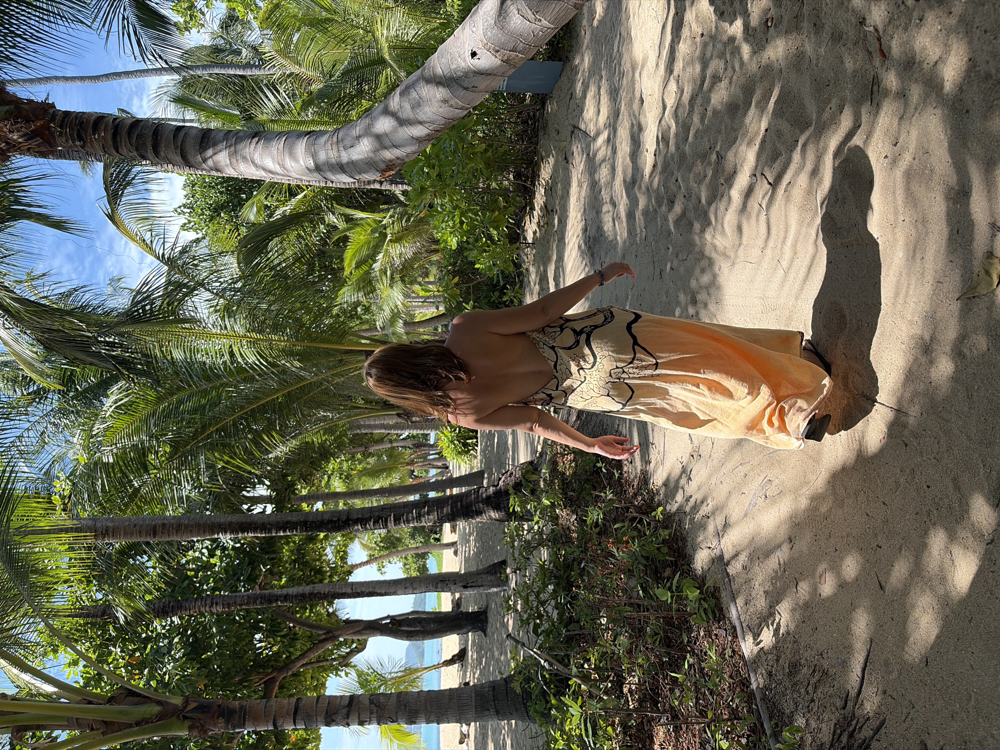 A woman in a long, sleeveless, flowing orange dress with a black pattern walks away from the camera on a sandy path lined with palm trees. The path is partially shaded by the palm fronds. In the background, a sandy beach with scattered lounge chairs and a body of turquoise water are visible. The sky is bright blue with some clouds. The location is identifiable as a beach on Ko Samui District, Thailand, due to the presence of tropical vegetation and the specific style of palm trees.