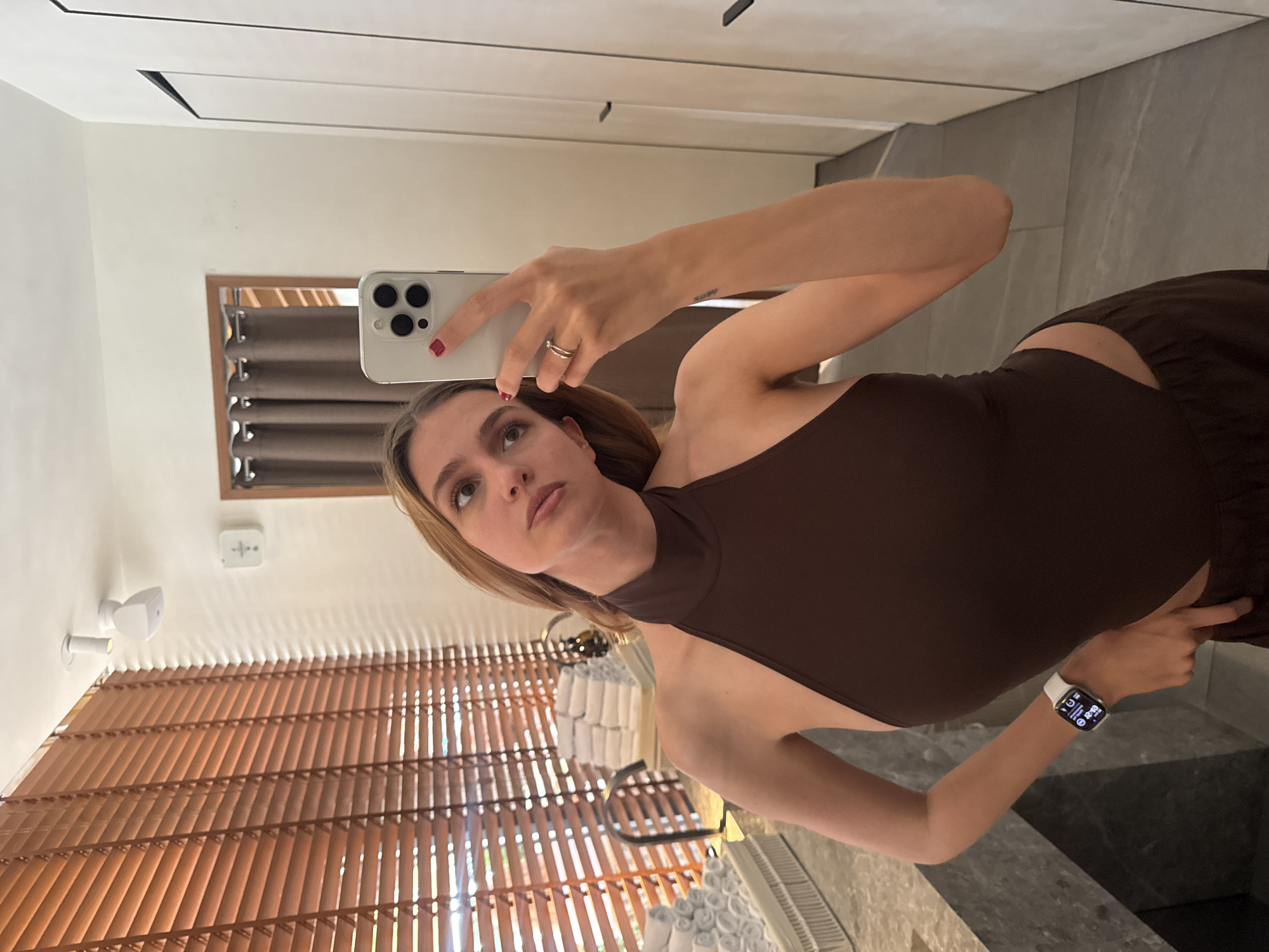 A fair-skinned woman is taking a selfie with a white smartphone. She is wearing a brown, high-necked top and is looking slightly up and to her left. The camera is tilted, showing a room with blinds, a window with curtains, a white ceiling fixture, and a marble countertop. Based on the angle and the items visible, the location is likely an apartment or hotel room, but the city of Yerevan, Armenia cannot be definitively confirmed without further details.