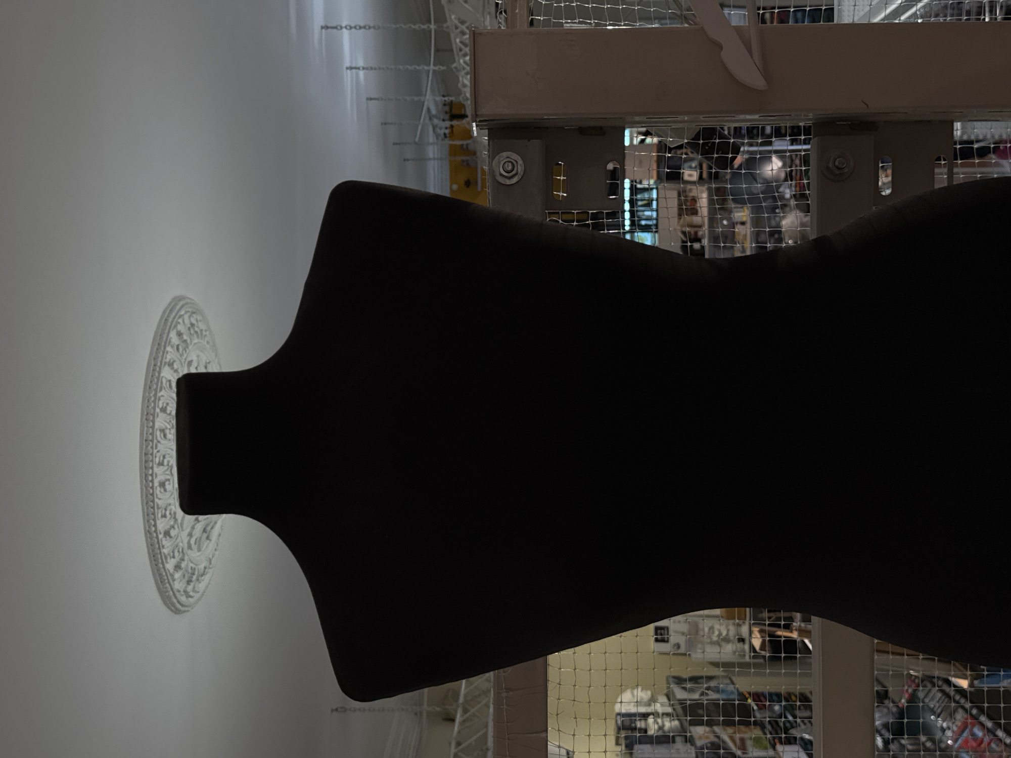 A dark, silhouetted mannequin is positioned in the foreground, facing away from the camera. The mannequin is dark and appears to be made of a matte material. The background reveals a store interior, with shelves filled with merchandise. The scene is brightly lit from above, casting a shadow of the mannequin onto the surrounding area. The location is not specified.