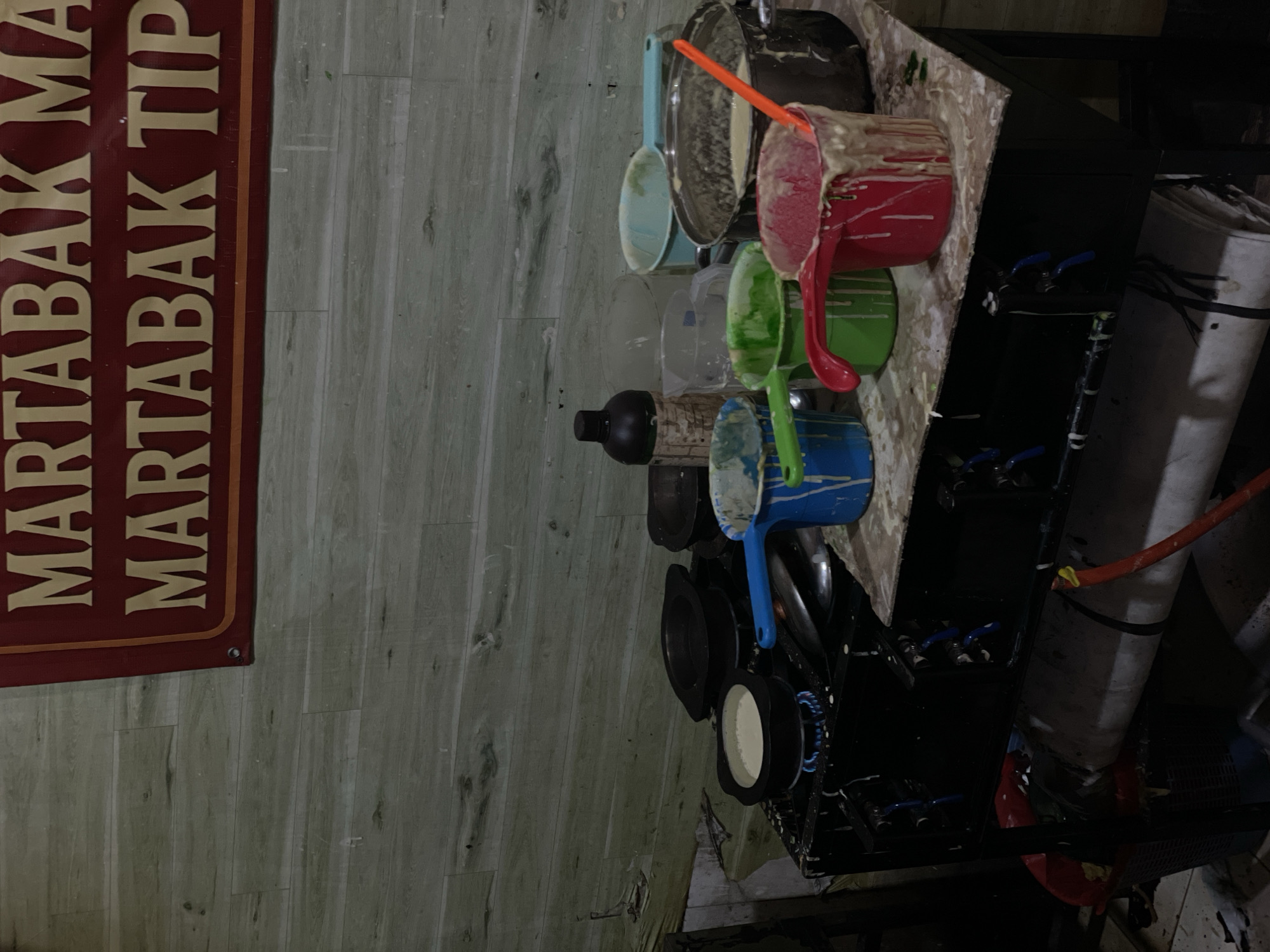This image shows a kitchen setup in North Jakarta, Indonesia. A vertical sign on the left reads "MARTABAK MA" and "MARTABAK TIP," indicating this is a food stall specializing in martabak, a popular street food. The setup includes several bowls, pots, and containers stacked on a counter with spatulas inserted in some of them. They appear to be various colors and contain mixtures that suggest they are ingredients used in the martabak preparation. Below the counter, there is a gas stove or cooking apparatus, with a gas cylinder beside it, connected by a red hose. The general environment appears to be a basic, informal cooking area, likely a street food vendor stall. The background wall is finished with imitation wood paneling.