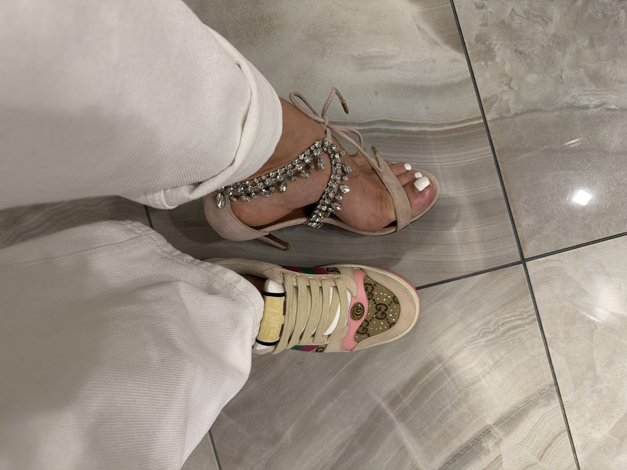 The image shows a person wearing white trousers, with one foot in a high-heeled sandal with crystal embellishments and the other in a Gucci sneaker. The person is standing on a tiled floor. The city and state are not identifiable.