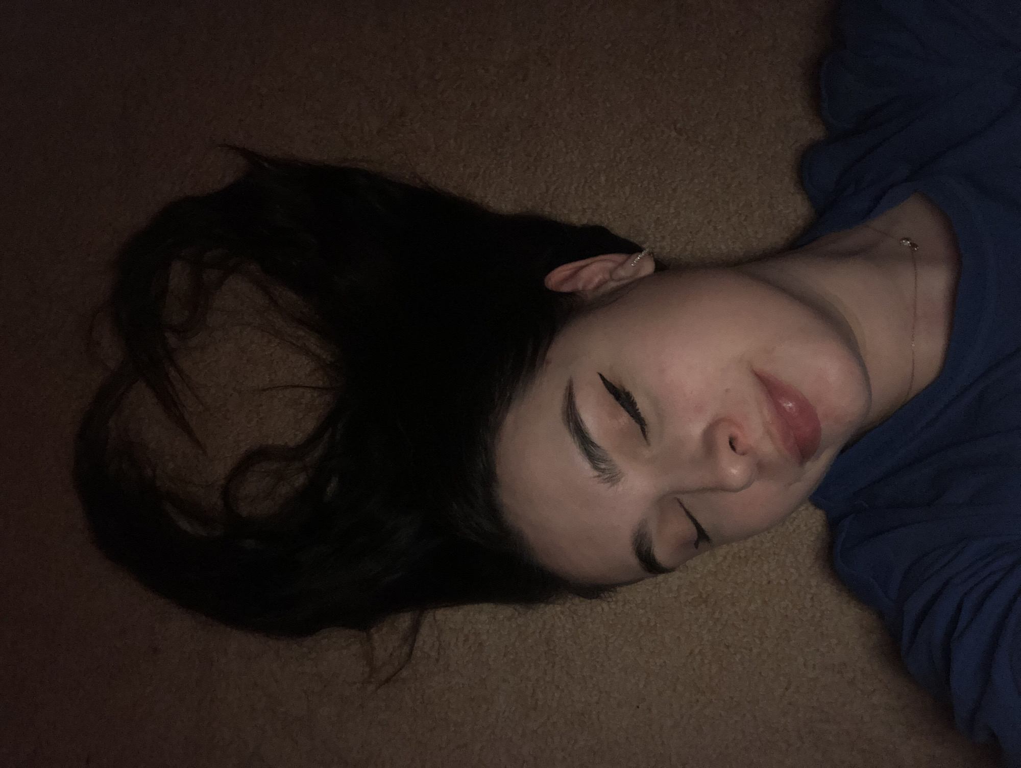 A young woman with dark hair is lying on a carpeted floor. Her hair is spread out beside her head. She is wearing a blue top and a delicate necklace. Her eyes are closed. The lighting is dim. The location is unknown; there is no information in the image to indicate it is Noviy, Russia, or any specific city.