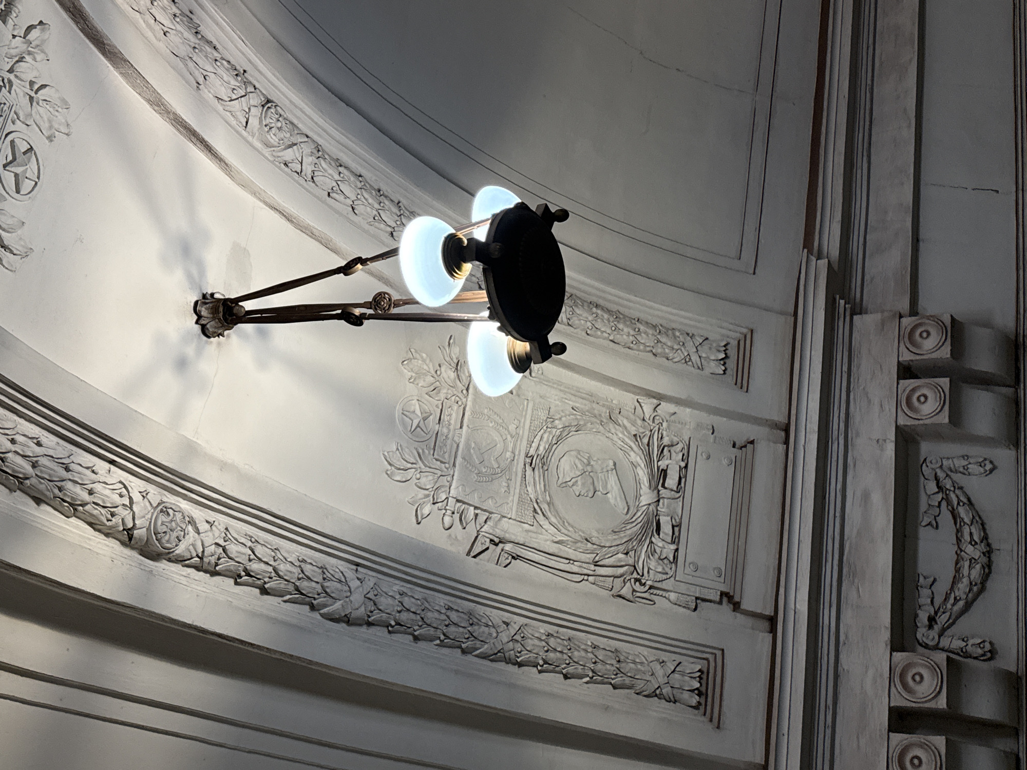 The image shows a view of the ceiling of what appears to be a grand interior space, possibly a subway station or a public building, with ornate plasterwork and a chandelier. The ceiling is arched and features intricate decorative moldings. Along the curve of the arch, a repeating pattern of laurel leaves is visible, interspersed with circular medallions.
In the center of the frame, a dark-colored chandelier hangs from the ceiling. It has a central circular base from which three arms extend, each supporting a frosted glass globe emitting a bright, cool-toned light. The chandelier's metalwork appears to be dark bronze or iron, with decorative elements at the points where the arms connect to the central body and at the top where it attaches to the ceiling.
To the left of the chandelier, a more elaborate decorative panel is mounted on the ceiling. This panel contains a relief sculpture depicting a profile bust of a person wearing a helmet, enclosed within a circular frame. Above the bust, a shield-like emblem is visible, featuring a hammer and sickle symbol, along with a star and floral motifs. This iconography suggests a connection to Soviet-era design or symbolism.
Below the arched ceiling, a horizontal band of decorative molding is present, featuring a relief of a garland of leaves and ribbons, flanked by circular elements. The overall impression is one of grandeur and historical significance, with a focus on architectural detail and decorative artistry. The lighting from the chandelier creates soft shadows on the white plaster surfaces. The specific location is unknown, but the style suggests a public monument or a building with significant historical or political importance, likely from the mid-20th century.