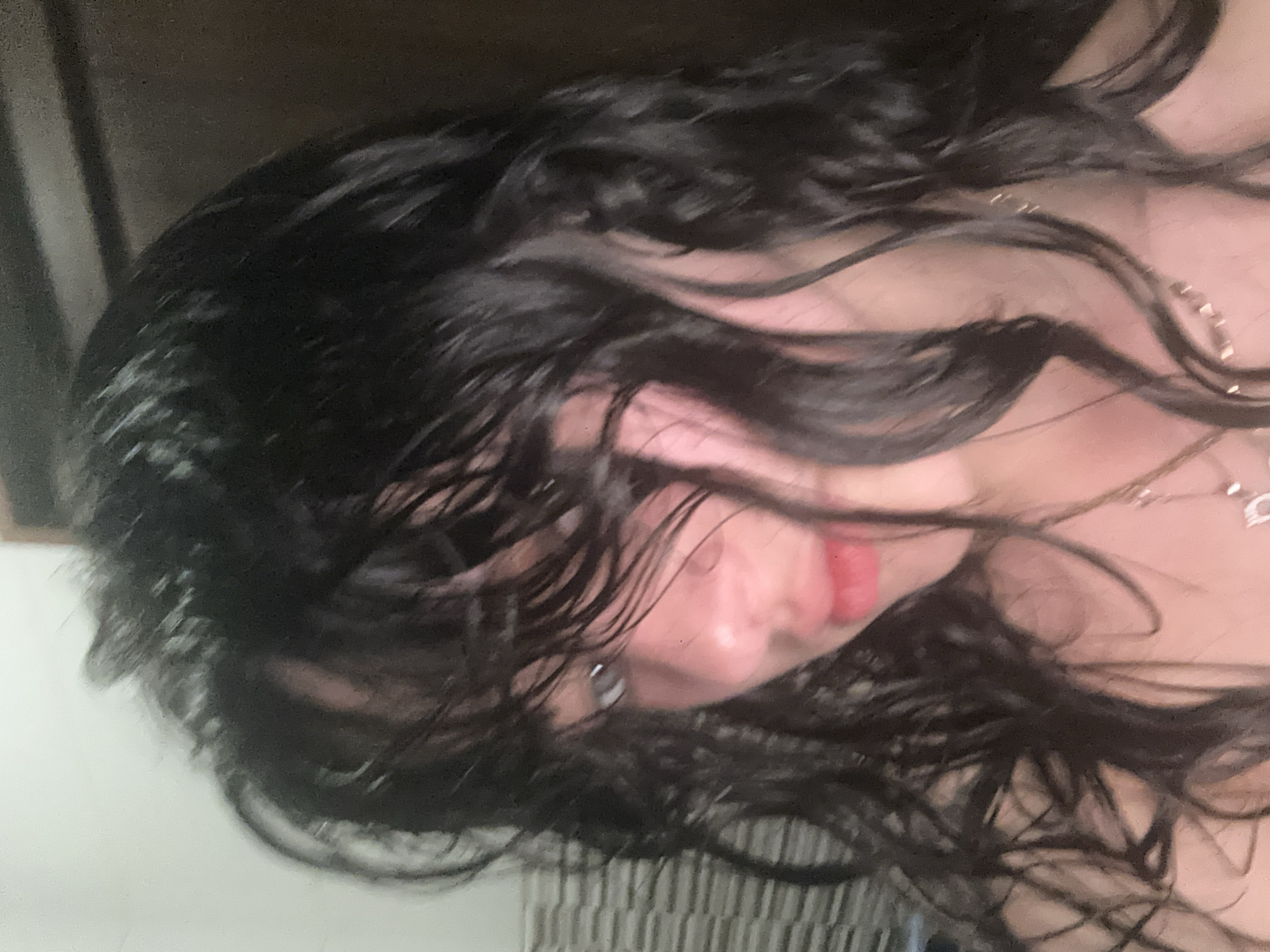 An image features a person with dark, wavy hair framing their face. They have fair skin, and their lips are visible with a red tint. A small piercing is located on the ear. A delicate necklace adorns their neck. The background appears to be a mix of light and dark tones, including furniture and possibly part of a wall. The location of this image cannot be identified as being in Yerevan, Armenia.