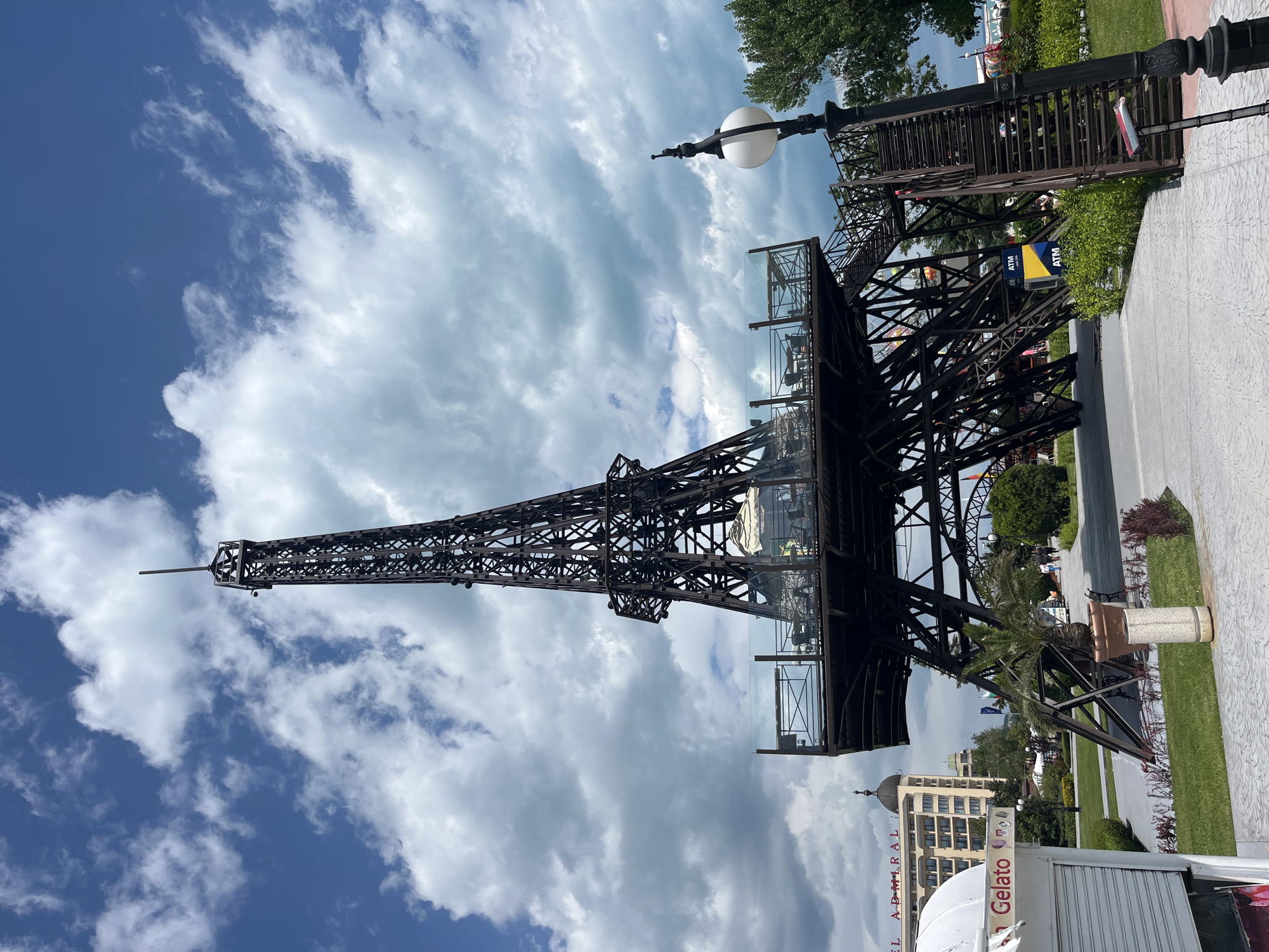 The image depicts a replica of the Eiffel Tower situated in Varna, Bulgaria. The structure is made of metal and appears to have glass-enclosed levels. The sky is visible behind the tower, filled with clouds and patches of blue. Below the tower, there are elements of a park or recreational area with greenery. In the foreground, parts of a building labeled "Gelato" and the "El Admiral" hotel can be observed. The area includes a street lamp and a paved pathway. An ATM is visible along with an observation tower.