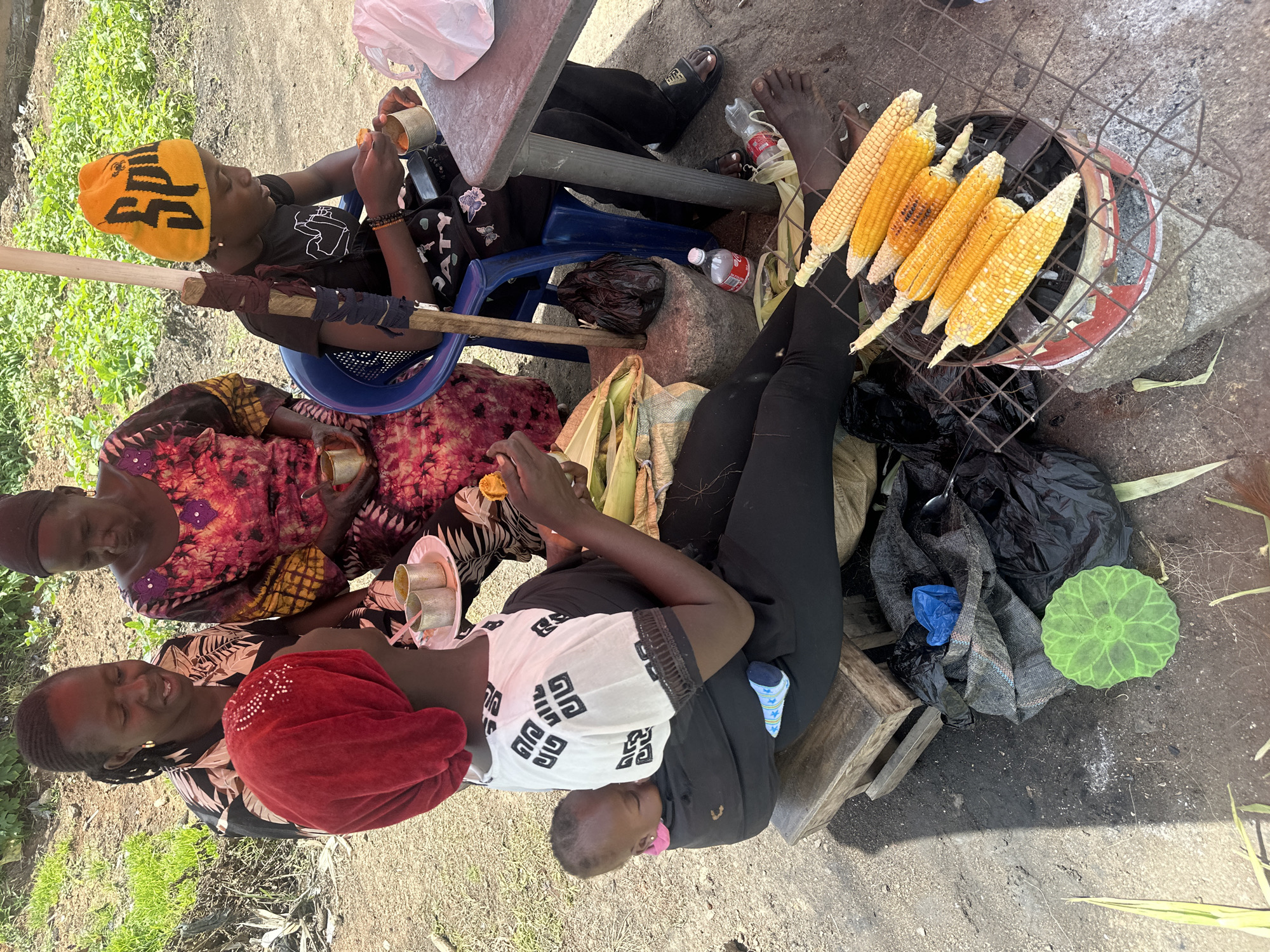 This media file captures an outdoor scene in Gura Top, Nigeria, likely during daylight hours as indicated by the bright lighting and shadows. The image features several individuals, appearing to be women and children, gathered around a small charcoal grill where corn is being roasted. One person, wearing an orange and black beanie, is seated in a blue chair, holding a small cup. Another individual, adorned in a colorful patterned dress, is also seated, holding a similar cup. A third person, wearing a white shirt with a black geometric pattern and a red headscarf, is actively engaged with the scene, possibly preparing food or interacting with others. The overall setting appears to be a casual, outdoor gathering, possibly a street vendor or a family event.
Visible objects include the charcoal grill with several ears of corn, plastic bags, a wooden crate, a plastic bottle, and plates with cups. The ground is a mix of dirt and what appears to be dry grass or vegetation. The presence of the grill and roasted corn suggests that food preparation and consumption is the primary activity. The expressions on the faces of the people, though partially obscured, suggest a relaxed and possibly joyful atmosphere. There is no visible text in the image. The scene is likely taking place in a rural or semi-urban environment, given the natural ground cover and the simple setup.