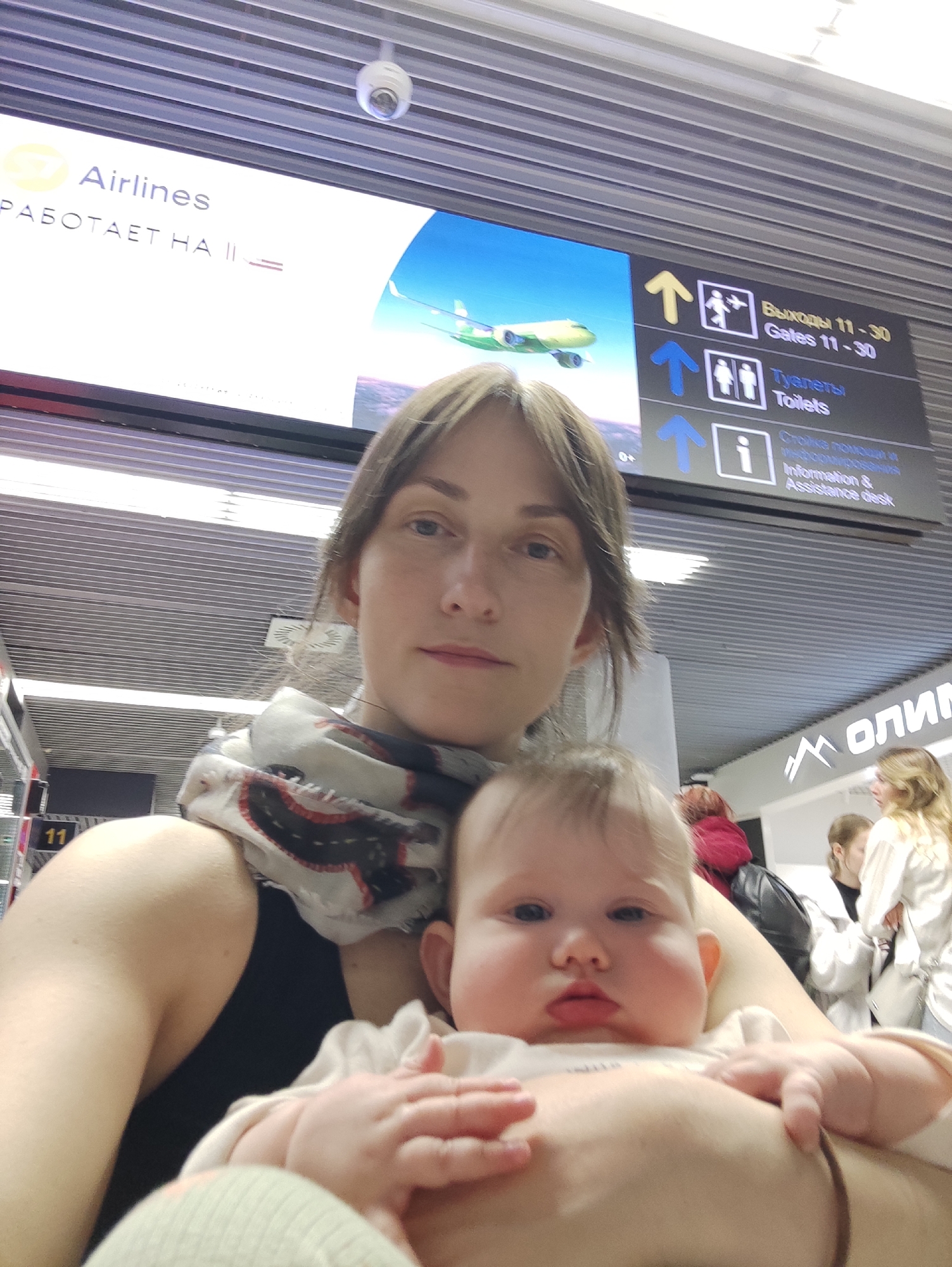 The image shows a woman holding a baby, likely in an airport, as suggested by the presence of signs indicating "Airlines," "Gates 11-30," "Toilets," and "Information & Assistance desk." The sign also indicates that the airline is operating on platform II. The presence of text in Russian (e.g., "Выходы," "Туалеты," "Стойка помощи") suggests the location is likely in Russia. The logo "ОЛИМП" is also visible. Given that "Олимп" is part of the name of the city of Sochi ("Сочи"), it can be inferred that this is Sochi International Airport, Russia. The woman is wearing a patterned scarf and a dark top. The baby is wearing a white top and has a focused expression. In the background, other people can be seen.