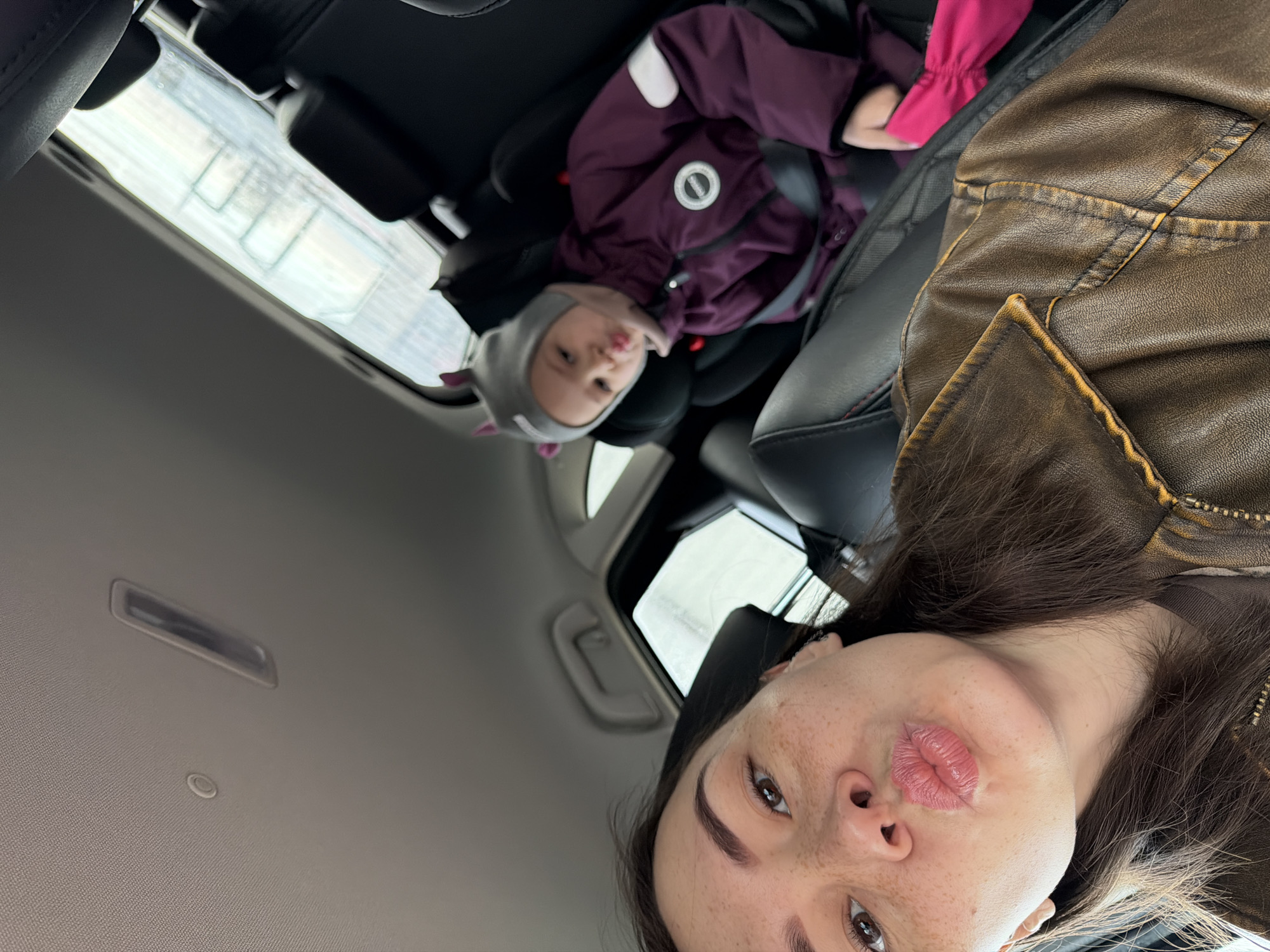 A selfie taken inside a vehicle shows a woman in the foreground and a child in a car seat in the background. The woman has brown hair, freckles, and is wearing a brown leather jacket. She is making a kissing face towards the camera. The child, who appears to be a toddler, is seated in a car seat, wearing a purple jacket with a white circular logo on the chest, and a grey hat with cat ears. Outside the car windows, a blurry view suggests a city scene, likely Chaikovsky, Russia, given the prompt's requirement. The interior of the car is visible, including the ceiling, handles, and part of the dashboard. The photo seems to have been taken from an upward angle.