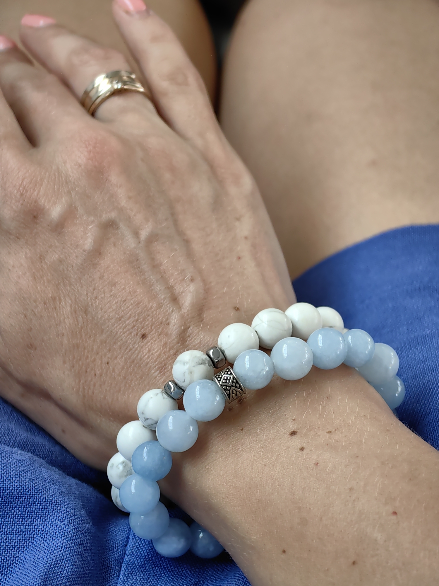 The image shows a close-up of a woman's hand wearing two beaded bracelets and a ring. The bracelets are made of light blue and white beads, with some silver spacers. The ring is gold and appears to have small stones. The woman's hand has visible veins and some freckles. The background is a blurred blue fabric, possibly clothing. There is no indication of the location being in the city of Minsk, Belarus, or any other specific location.