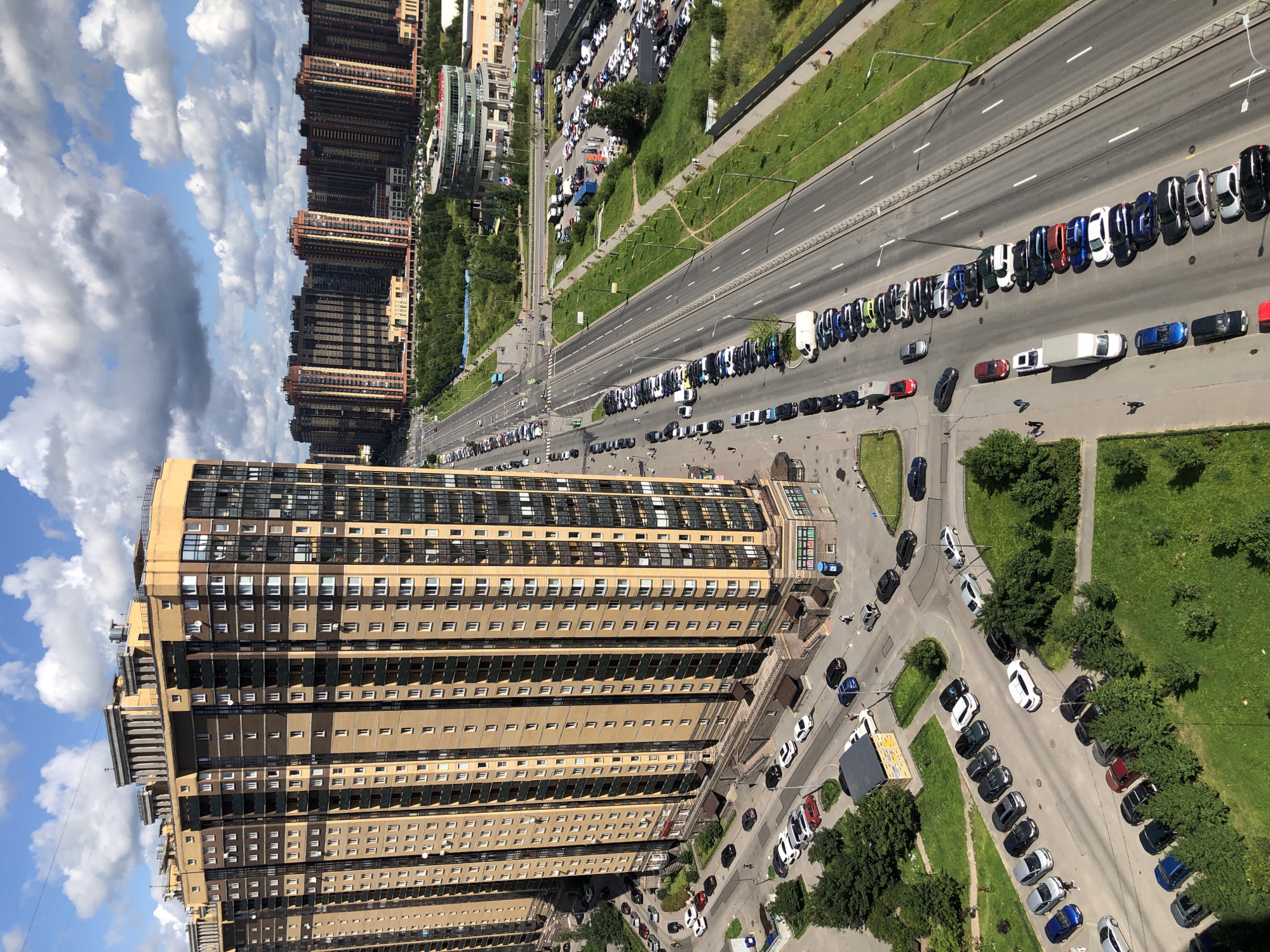 This aerial photograph depicts a high-rise apartment building in an urban environment. The building, with its beige and brown facade, features numerous windows and balconies. Adjacent to the building is a multi-lane road with a significant number of parked cars lining both sides. Beyond the road, more high-rise buildings and green spaces are visible. The sky is partly cloudy, with distinct white clouds against a blue backdrop. The overall scene suggests a residential area within a city.